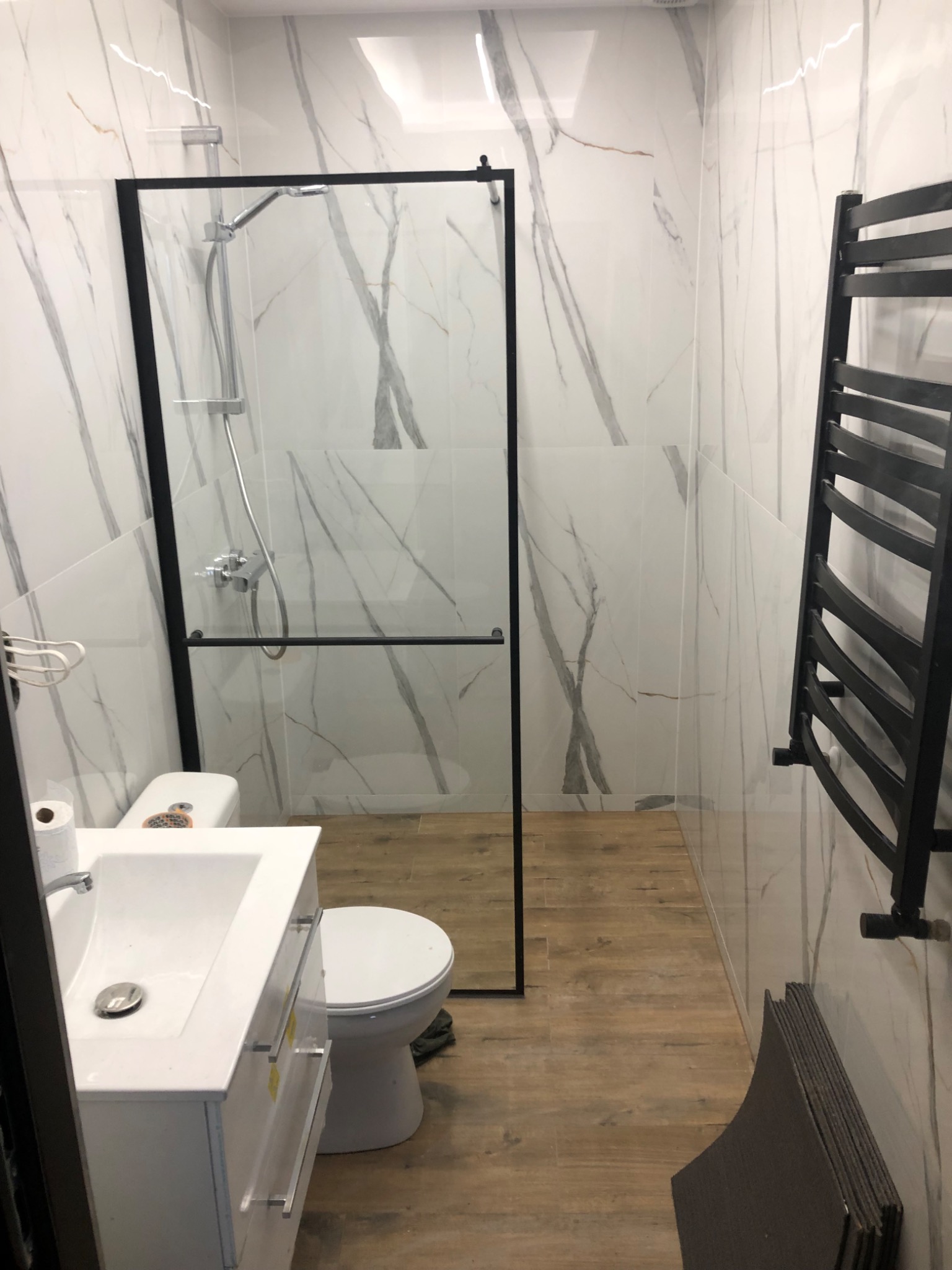 Nowoczesna łazienka z kabiną prysznicową typu walk-in, umywalką z szafką, toaletą i grzejnikiem drabinkowym w kolorze czarnym na tle płytek imitujących marmur.