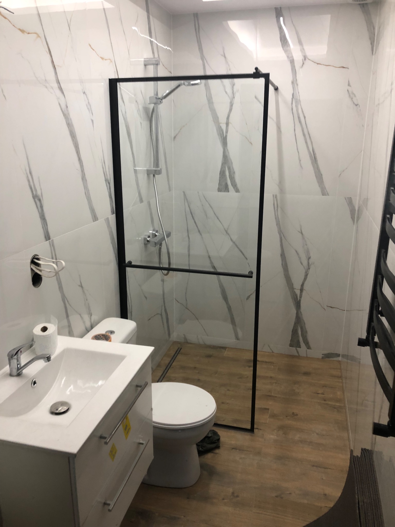 Nowoczesna łazienka z kabiną prysznicową typu walk-in, białą umywalką z szafką i toaletą, wykończona płytkami imitującymi marmur oraz drewnopodobną podłogą.