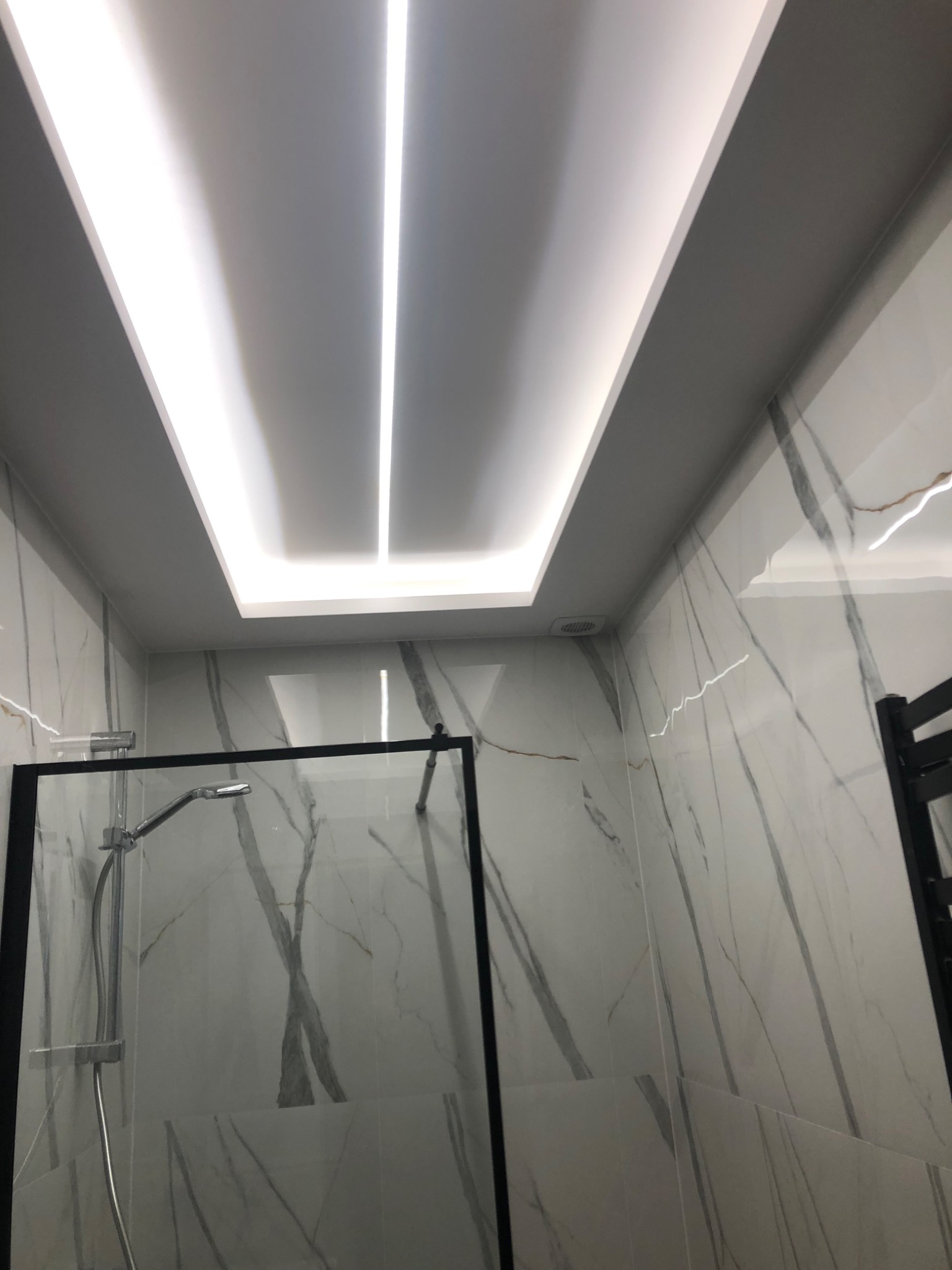 Nowoczesna łazienka z kabiną prysznicową typu walk-in, wykończona płytkami imitującymi marmur i oświetleniem LED w suficie podwieszanym.