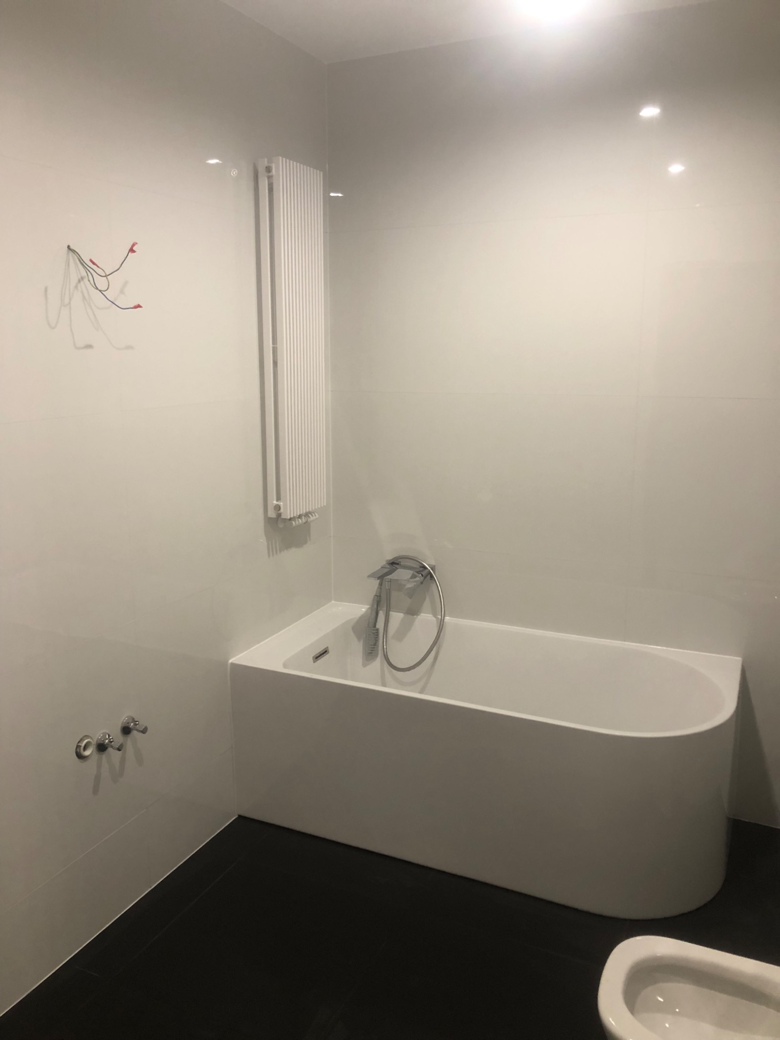 Minimalistyczna łazienka z białą wanną prostokątną, grzejnikiem drabinkowym na ścianie wyłożonej białymi płytkami oraz czarną podłogą z płytek. Na ścianie widoczne ślady po instalacji elektrycznej.