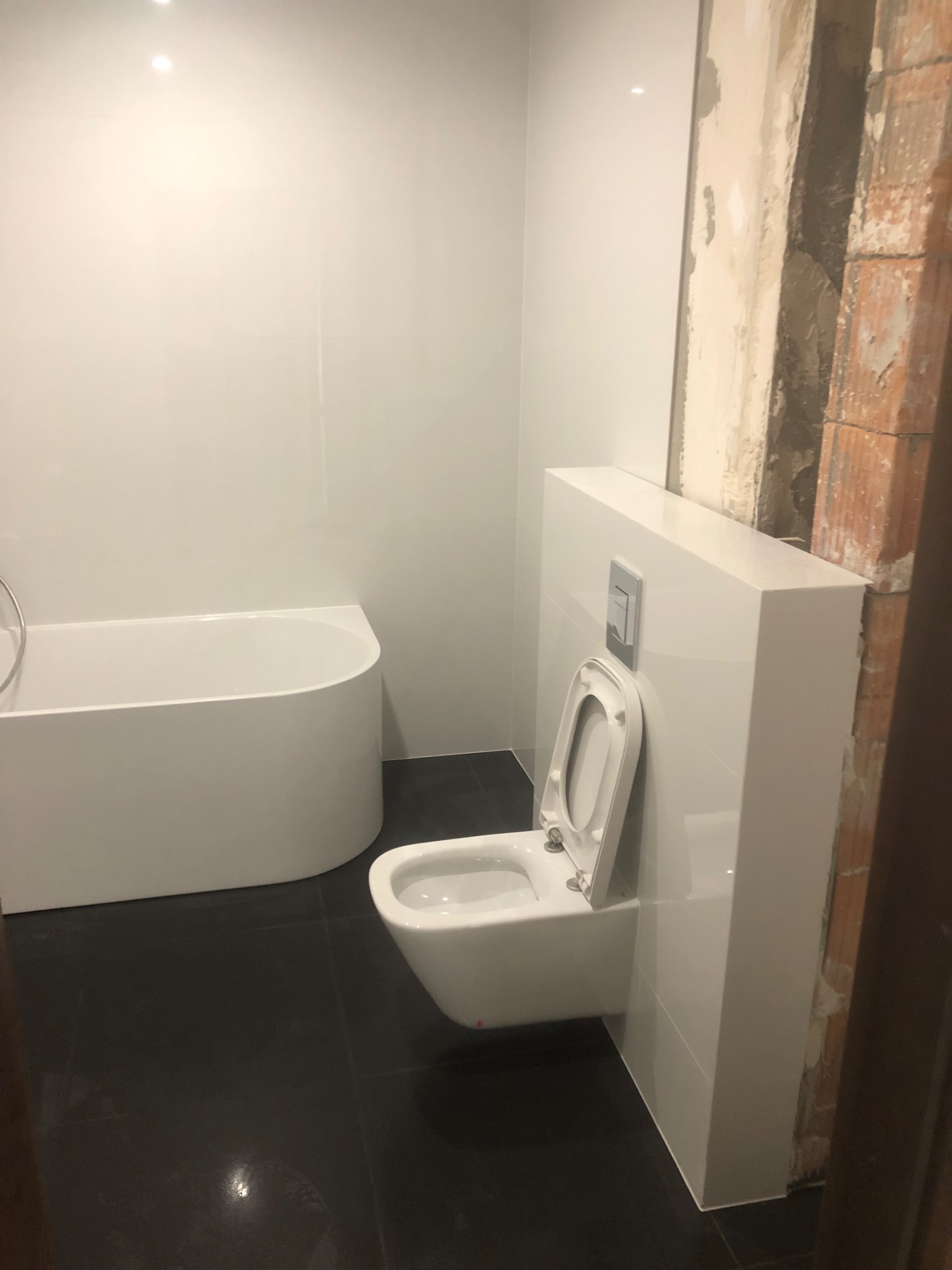 Wnętrze łazienki w trakcie prac wykończeniowych: biała wanna, podwieszana toaleta z zabudową, czarna podłoga, ściana w trakcie obróbki przy toalecie.