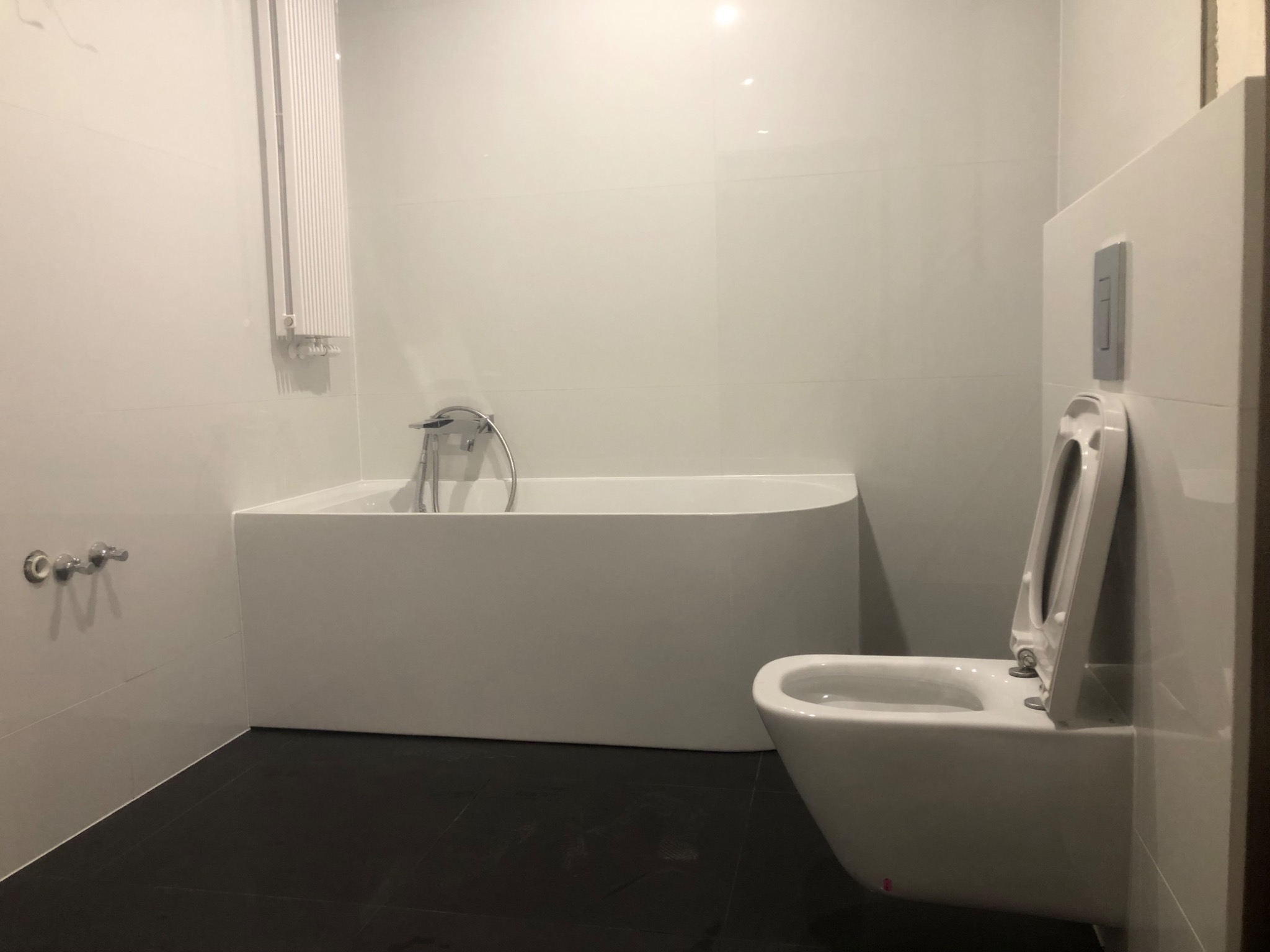 Minimalistyczna, nowoczesna łazienka z białą wanną, podwieszaną toaletą z szarą płytką przycisku spłukiwania i czarną podłogą, oświetlona światłem odbijającym się od białych płytek ściennych.