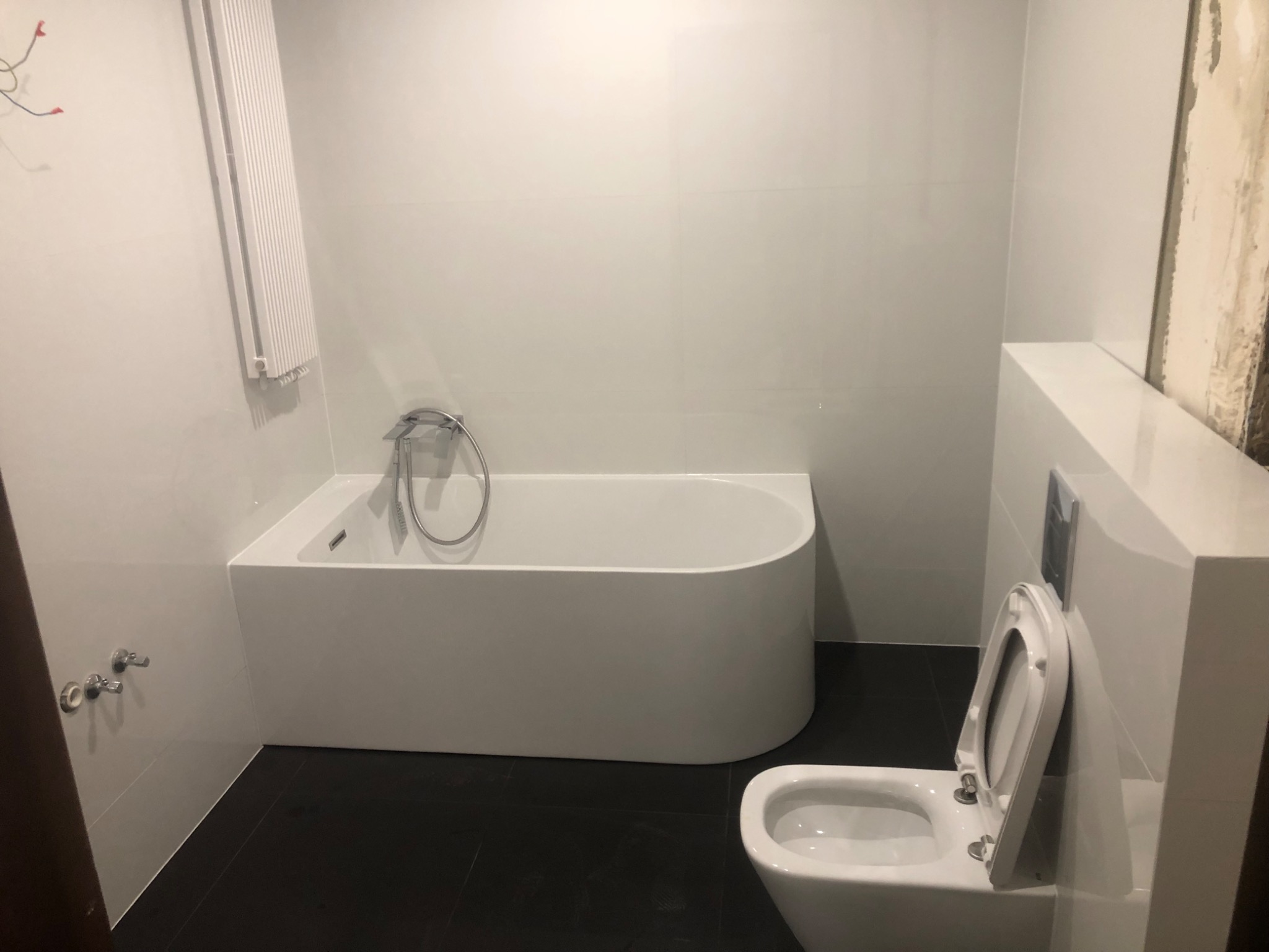 Nowoczesna łazienka z białą wanną, podwieszaną toaletą i ciemną podłogą z płytek, grzejnik drabinkowy na ścianie, widoczne przyłącza wodne.