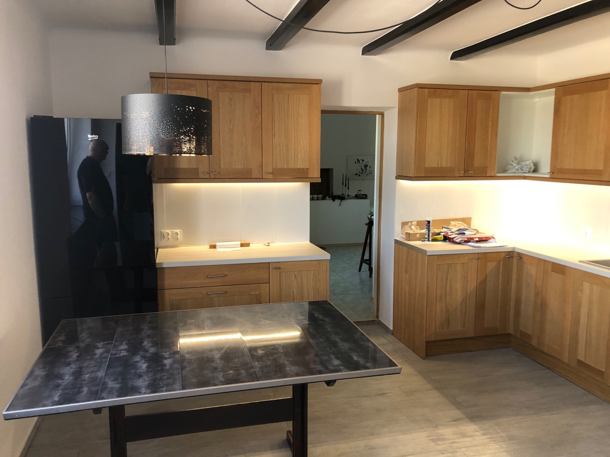 Jasna kuchnia z drewnianymi szafkami, czarnym blatem stołu z połyskiem i ozdobną lampą z ażurowym abażurem. Widoczny fragment czarnej lodówki i drewniane belki na suficie.