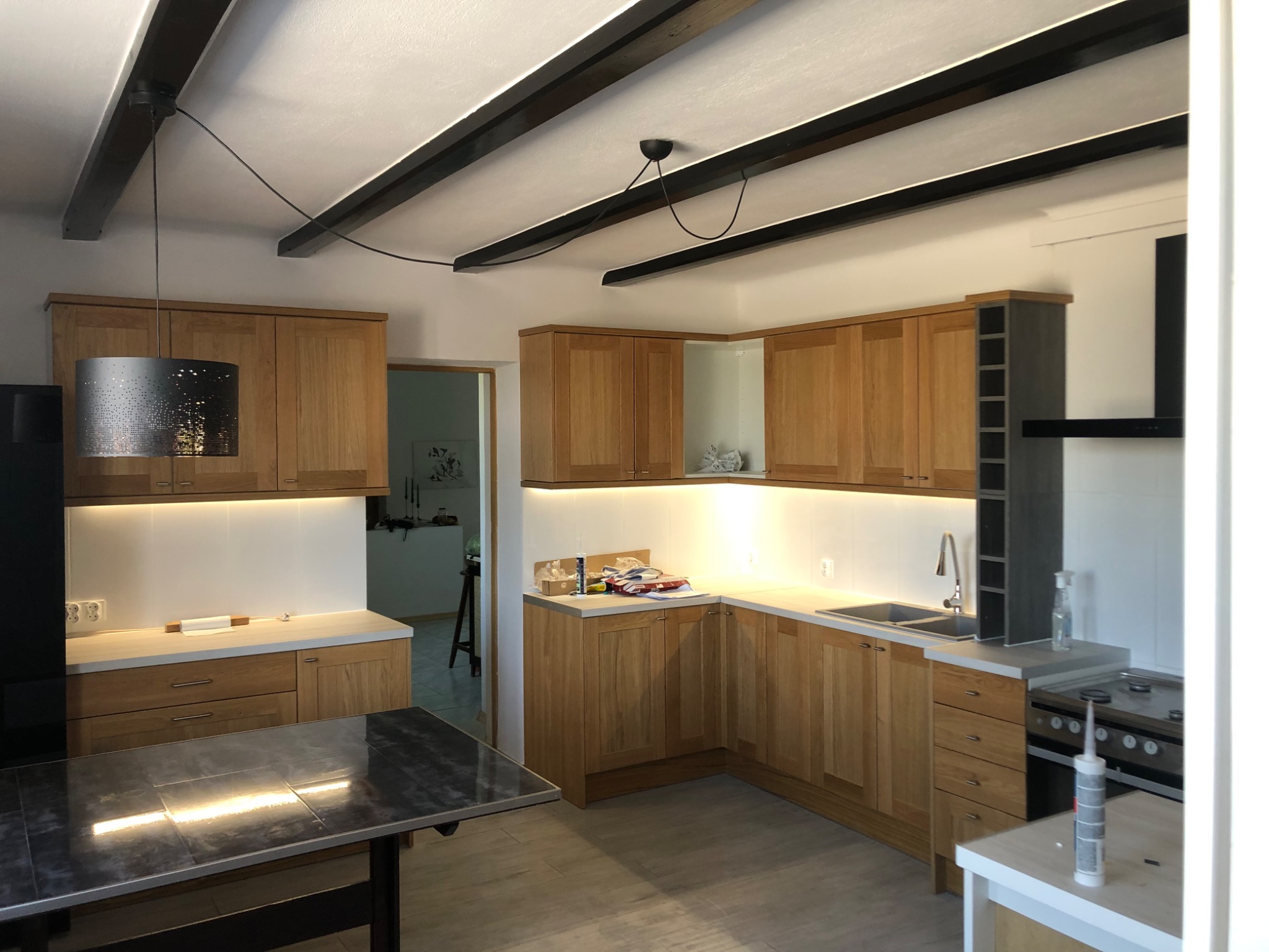 Jasna kuchnia z drewnianymi szafkami, białymi blatami i dekoracyjnymi belkami na suficie, oświetlona lampą z ażurowym abażurem i dodatkowym oświetleniem LED pod szafkami.
