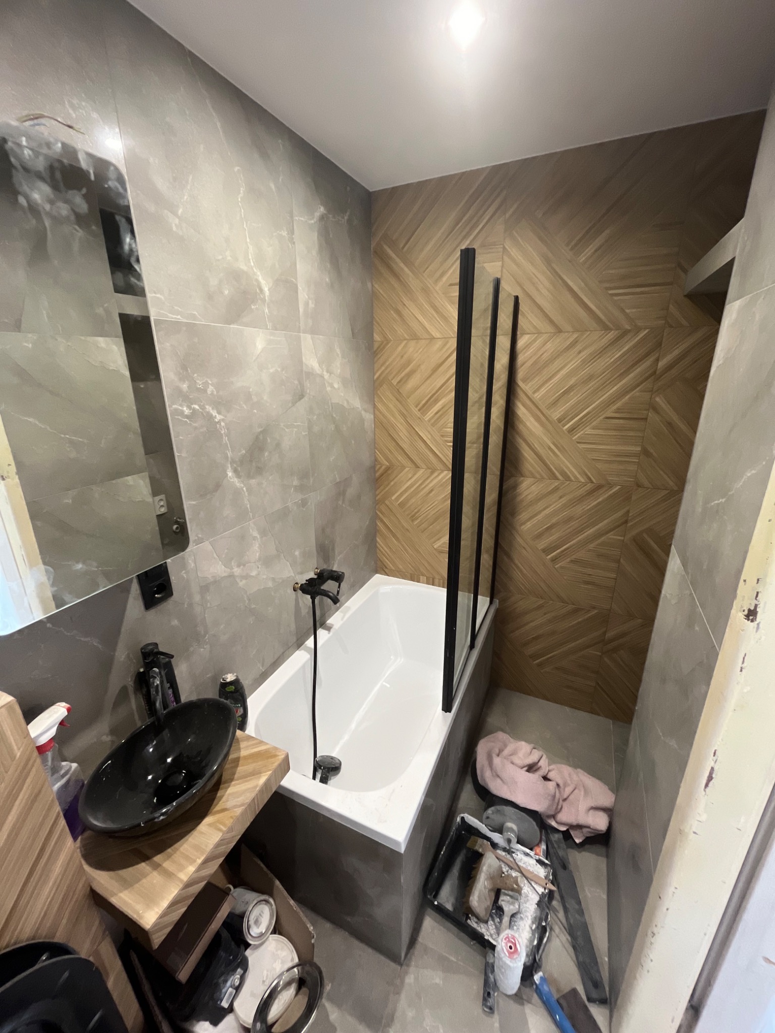 Nowoczesna łazienka w trakcie wykańczania: szare płytki imitujące marmur, drewnopodobna mozaika na jednej ze ścian, czarna umywalka nablatowa, biała wanna z czarną armaturą i szklanym parawanem...