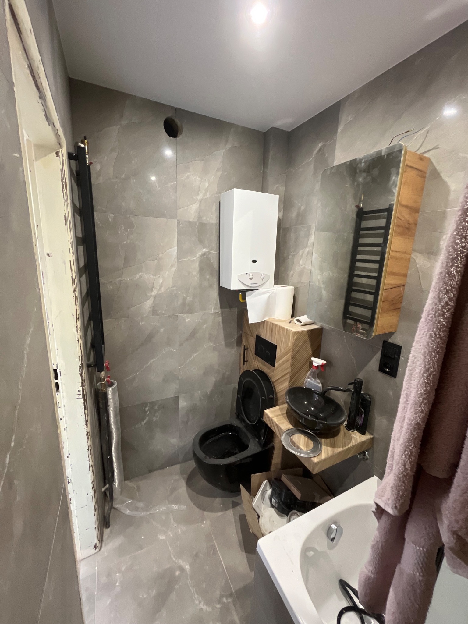 Łazienka w trakcie remontu: szare płytki imitujące marmur, wiszący grzejnik, umywalka nablatowa z drewnianą szafką, toaleta w zabudowie, wiszący piec gazowy. Widoczne narzędzia i akcesoria łazienkowe.