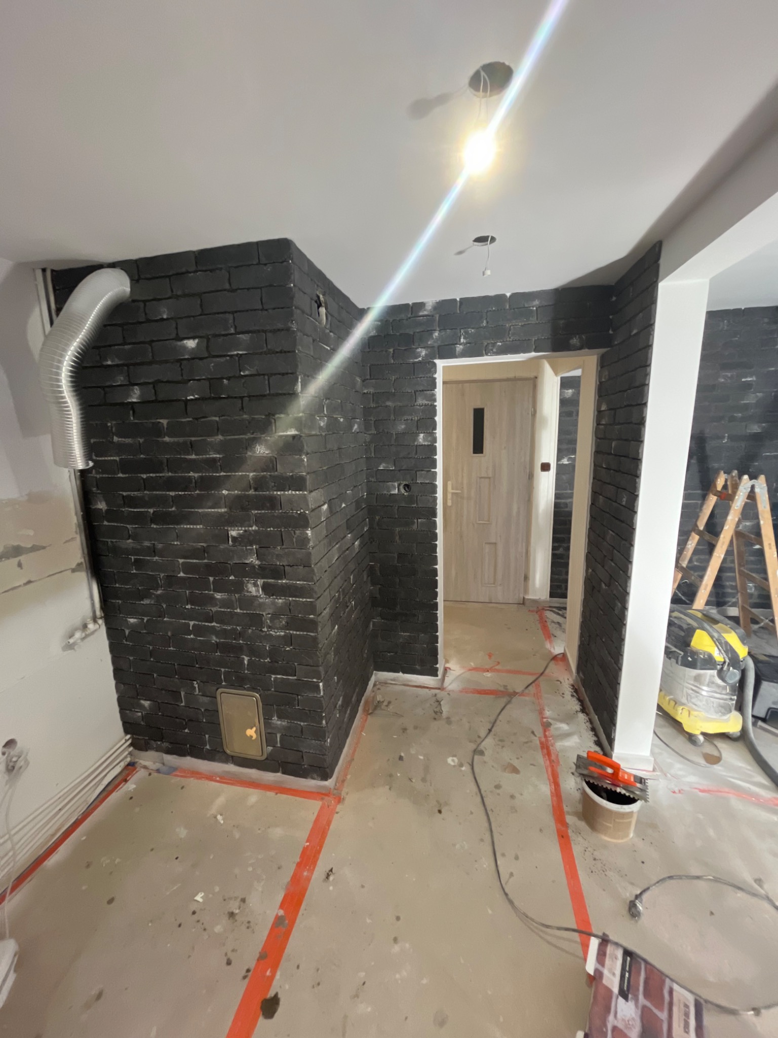 Wnętrze w trakcie wykańczania z pomalowaną na czarno cegłą na ścianach, widoczny odkurzacz przemysłowy, drabina, wiadro z narzędziami, taśma malarska na podłodze i rura wentylacyjna.