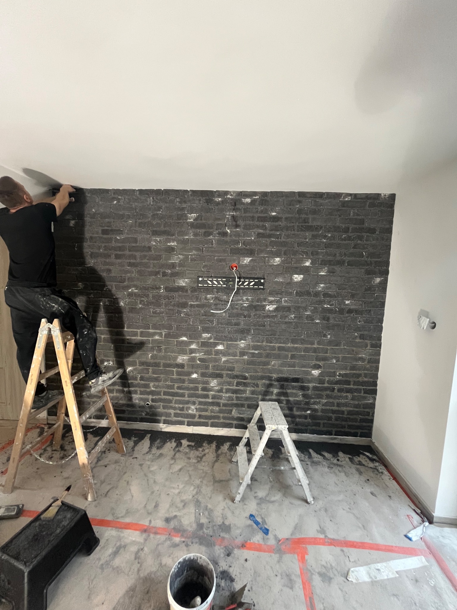 Mężczyzna na drabinie wygładza szpachlą ścianę z imitacją czarnej cegły z białymi przetarciami, widoczne narzędzia i akcesoria malarskie, podłoga zabezpieczona folią malarską.