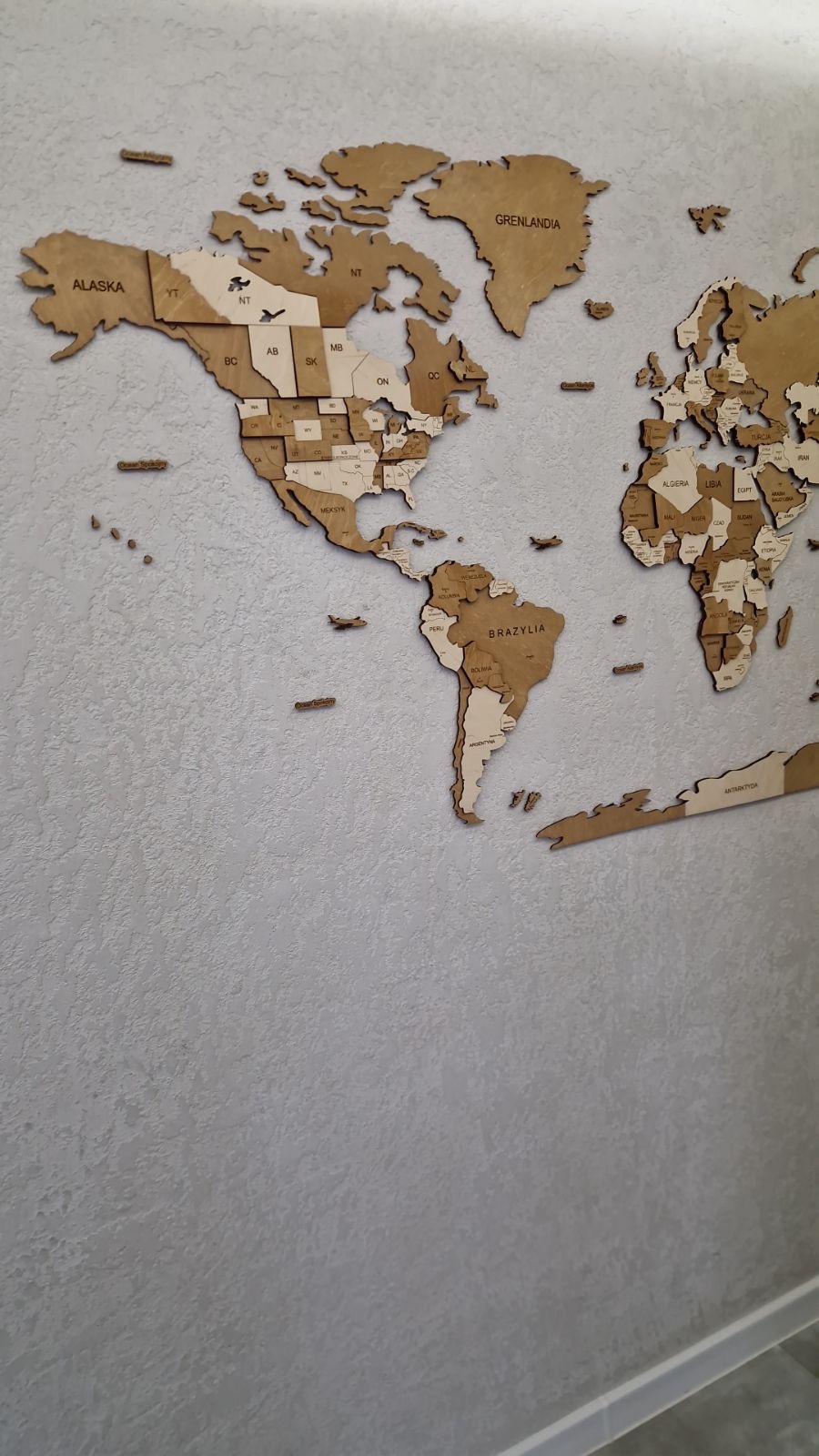 Dekoracyjna, drewniana mapa świata z grawerowanymi nazwami krajów i stanów USA, zamontowana na ścianie o delikatnej fakturze.