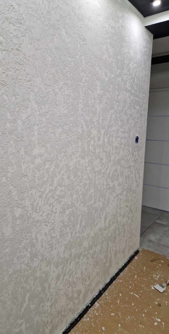 Ściana wewnętrzna z dekoracyjną, jasną strukturą, widoczny fragment podłogi z drobnymi zabrudzeniami i czarną listwą przypodłogową.