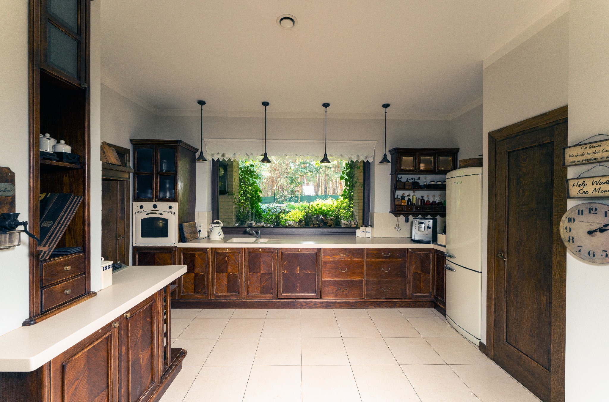 Rustykalna kuchnia z ciemnego drewna z dużym oknem wychodzącym na zieleń, biała lodówka retro i zegar na ścianie.