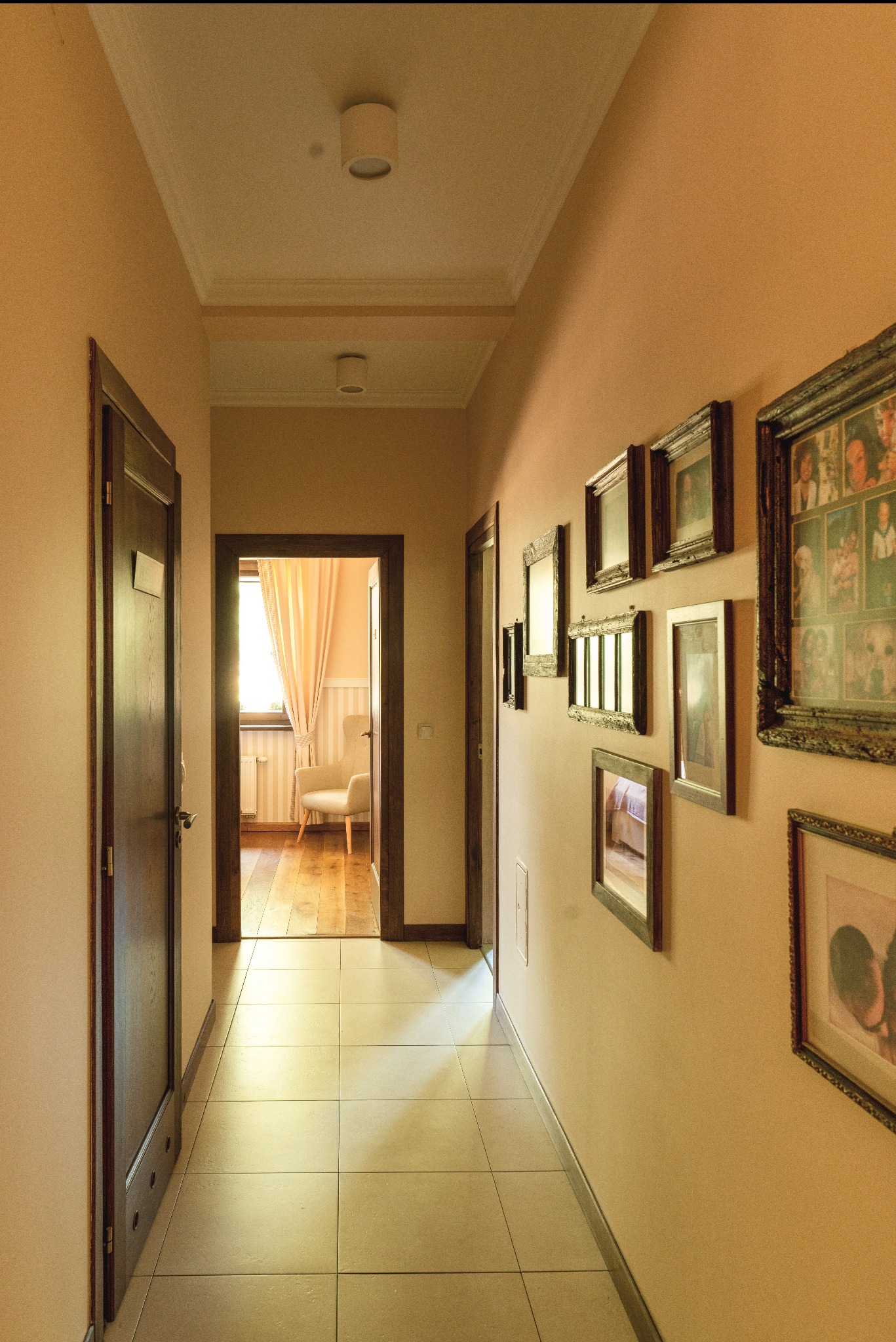 Korytarz w ciepłych odcieniach z serią ramek na ścianie, widok na jasny pokój z fotelem i zasłonami.