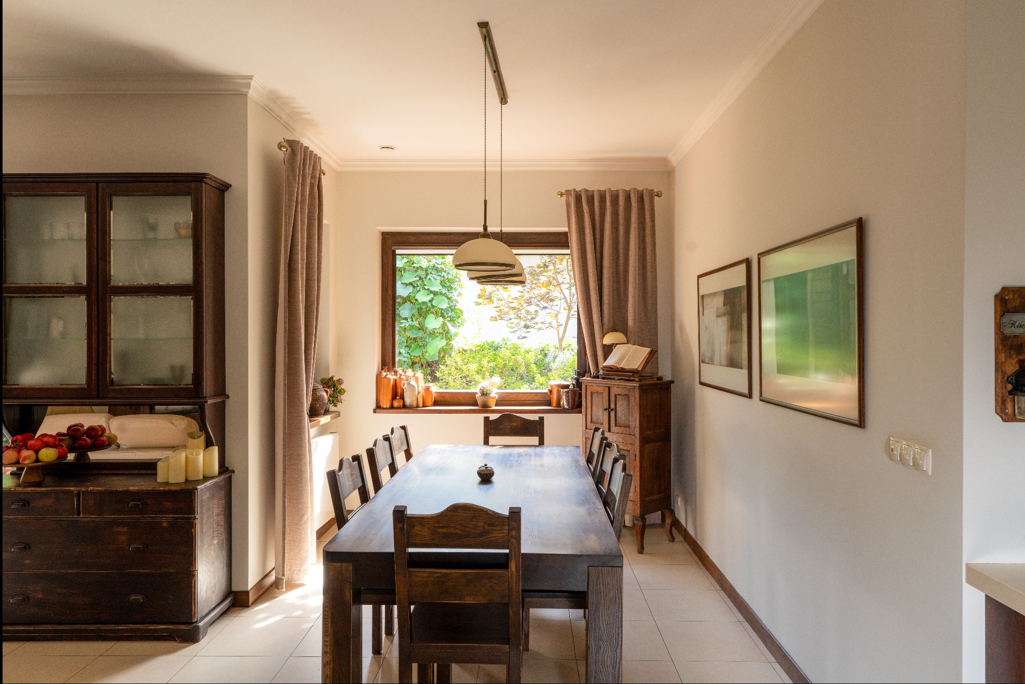Jasne wnętrze jadalni z dużym drewnianym stołem, krzesłami, kredensem z owocami i dekoracjami, okno z widokiem na zieleń, wisząca lampa i obrazy na ścianie.