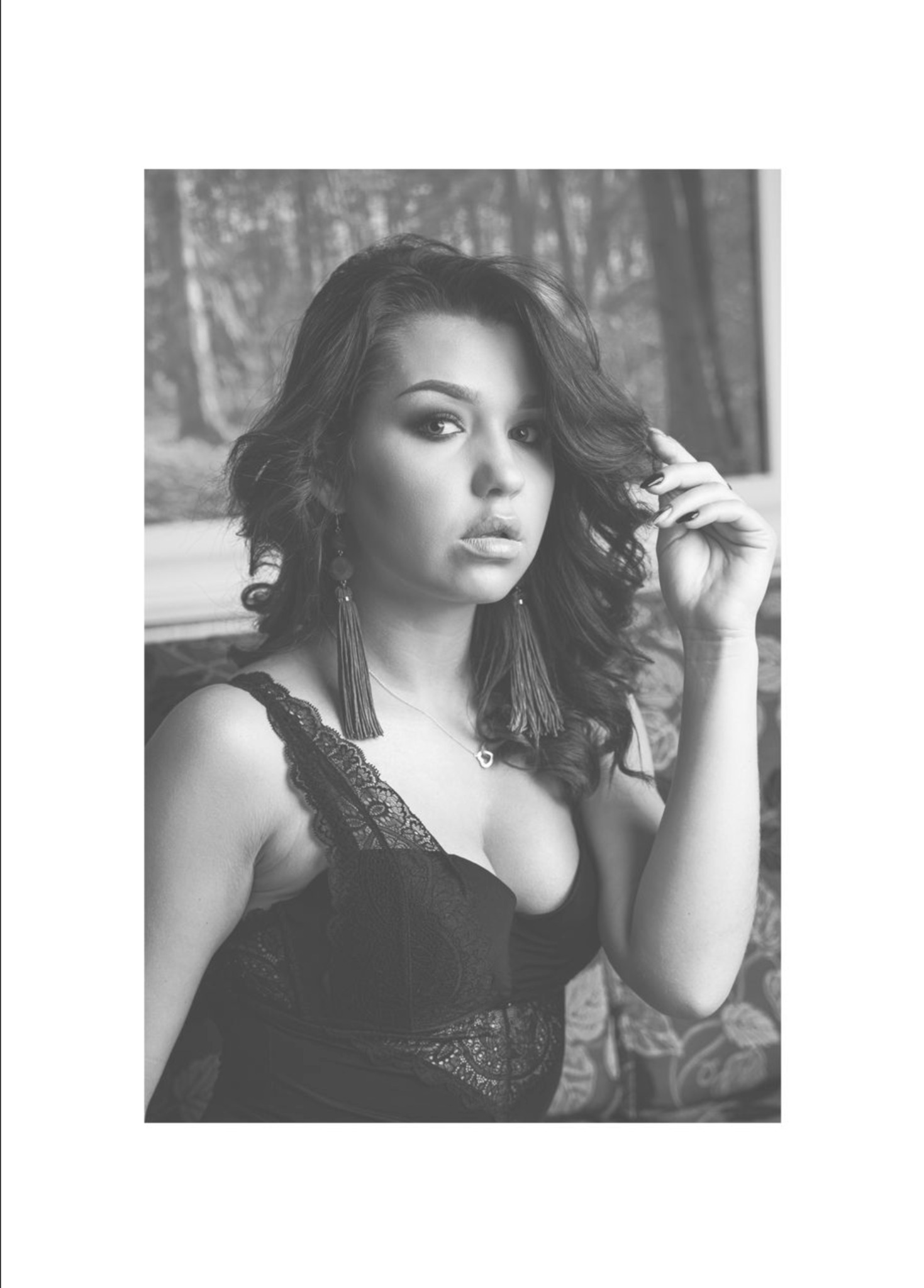 Portret kobiety w czarno-białej tonacji, z długimi kolczykami i delikatnym makijażem, pozującej na tle wzorzystej tapety imitującej las.