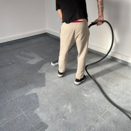 MK Clean Sp. z o.o. - Mężczyzna z tatuażami na ramieniu czyści szarą, kwadratową podłogę w pustym pomieszczeniu za pomocą odkurzacza piorącego.