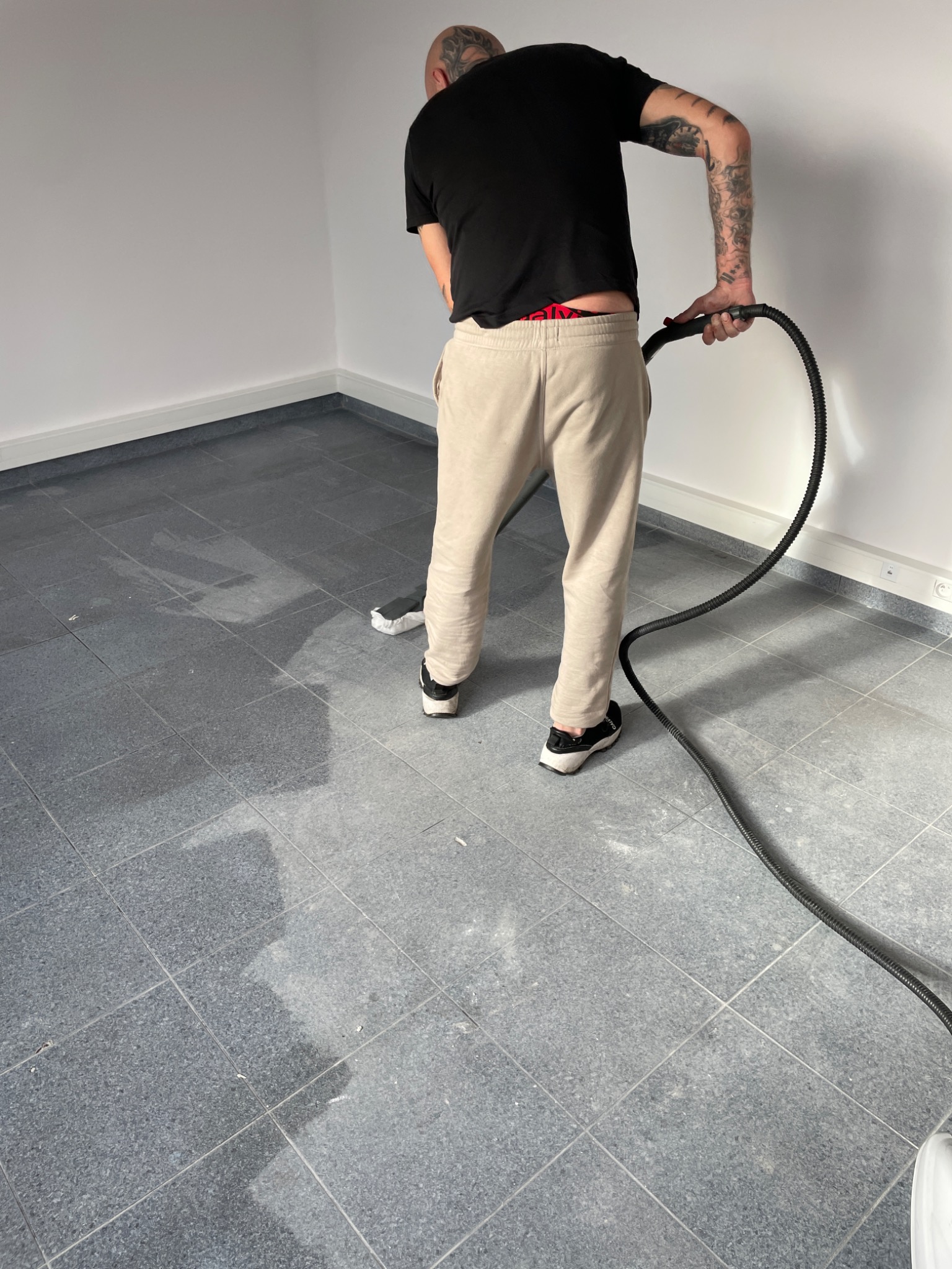 Mężczyzna z tatuażami na ramieniu czyści szarą, kwadratową podłogę w pustym pomieszczeniu za pomocą odkurzacza piorącego.