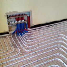 Instalacja ogrzewania podłogowego z rozdzielaczem w ścianie, widoczne białe rury układane na siatce z czerwonymi kwadratami, czarna izolacja przy ścianie.