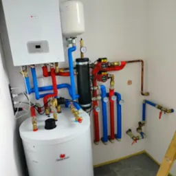 Kompletna instalacja grzewcza z piecem, zbiornikiem i rurami w kolorach czerwonym i niebieskim, widoczna drabina w rogu pomieszczenia.