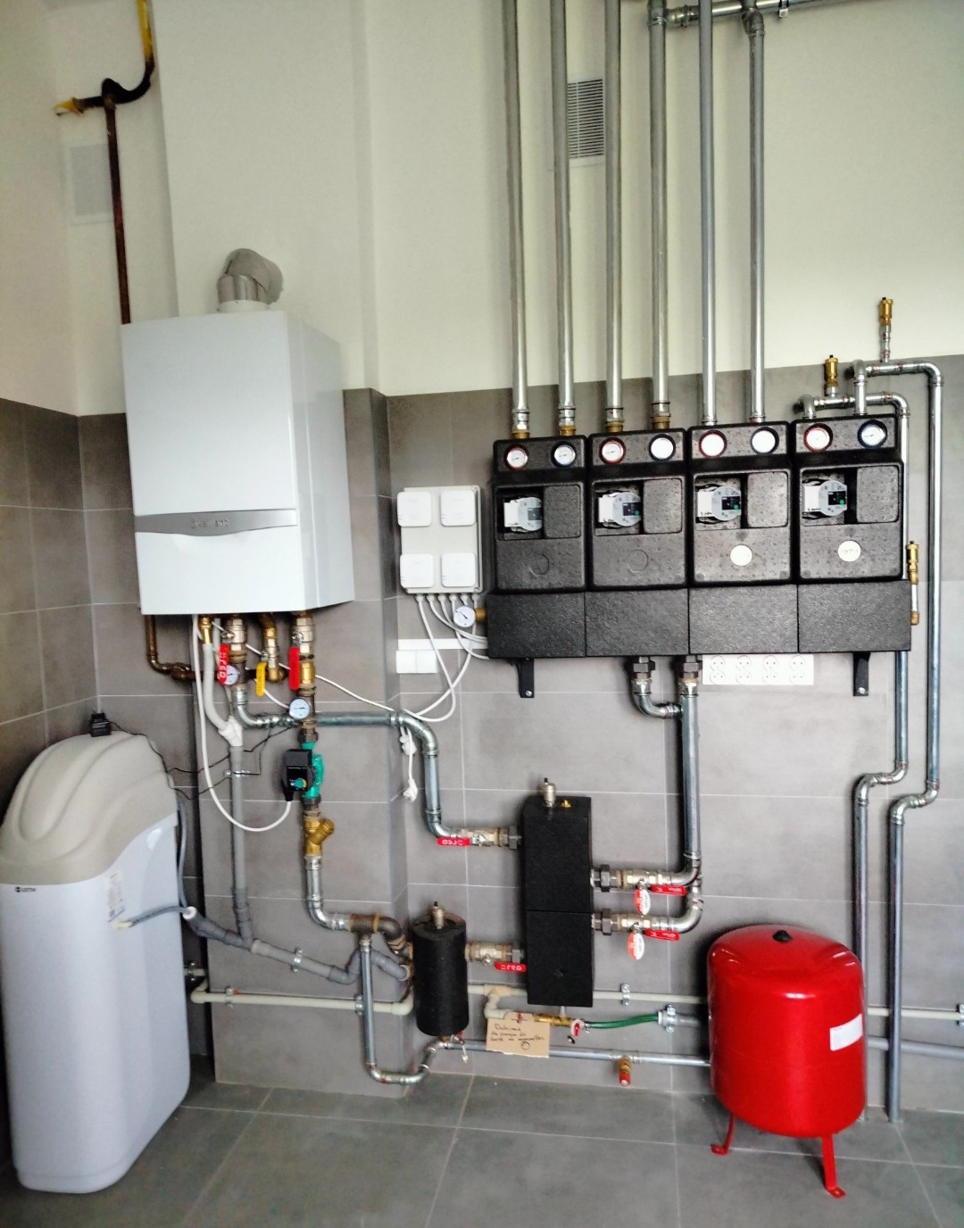 Nowoczesna kotłownia z białym piecem gazowym, stacją uzdatniania wody, rozdzielaczami grzewczymi i czerwonym zbiornikiem wyrównawczym na tle szarych płytek.