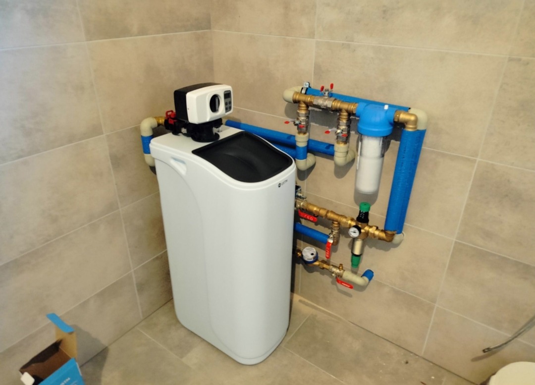 Nowoczesny zmiękczacz wody z elektronicznym sterowaniem, zainstalowany w rogu łazienki z beżowymi płytkami, widoczne rury i zawory doprowadzające wodę.