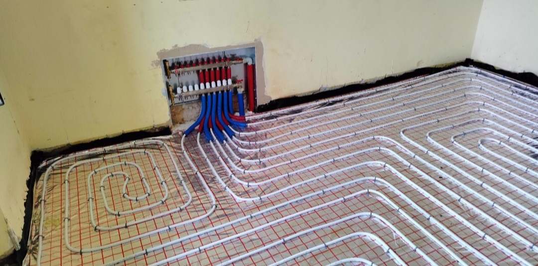 Instalacja ogrzewania podłogowego z rozdzielaczem w ścianie, widoczne białe rury układane na siatce z czerwonymi kwadratami, czarna izolacja przy ścianie.
