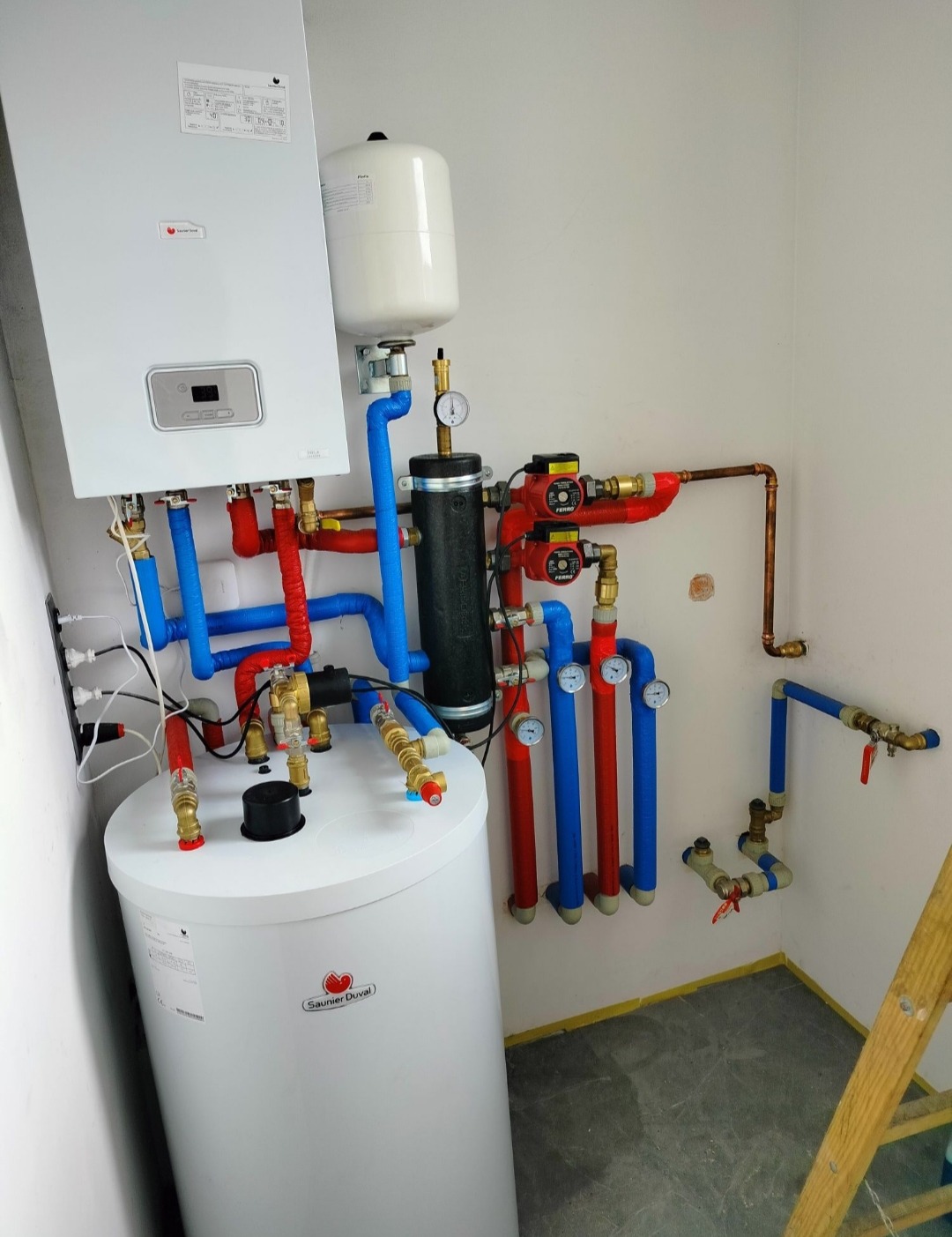 Kompletna instalacja grzewcza z piecem, zbiornikiem i rurami w kolorach czerwonym i niebieskim, widoczna drabina w rogu pomieszczenia.
