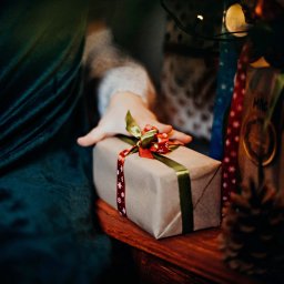 Agata Januszewska - Dłoń dziecka sięga po prezent zapakowany w szary papier i przewiązany zieloną oraz czerwoną wstążką w śnieżynki, leżący na drewnianym stole, w tle fragment zielonego aksamitnego materiału...