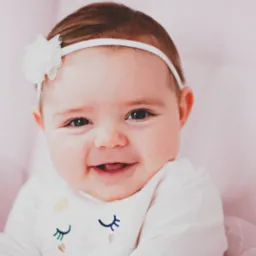 Uśmiechnięta niemowlęca dziewczynka z białym kwiatem na opasce, ubrana w białą bluzkę z haftowanymi oczami i złotymi elementami, na tle różowej poduszki.