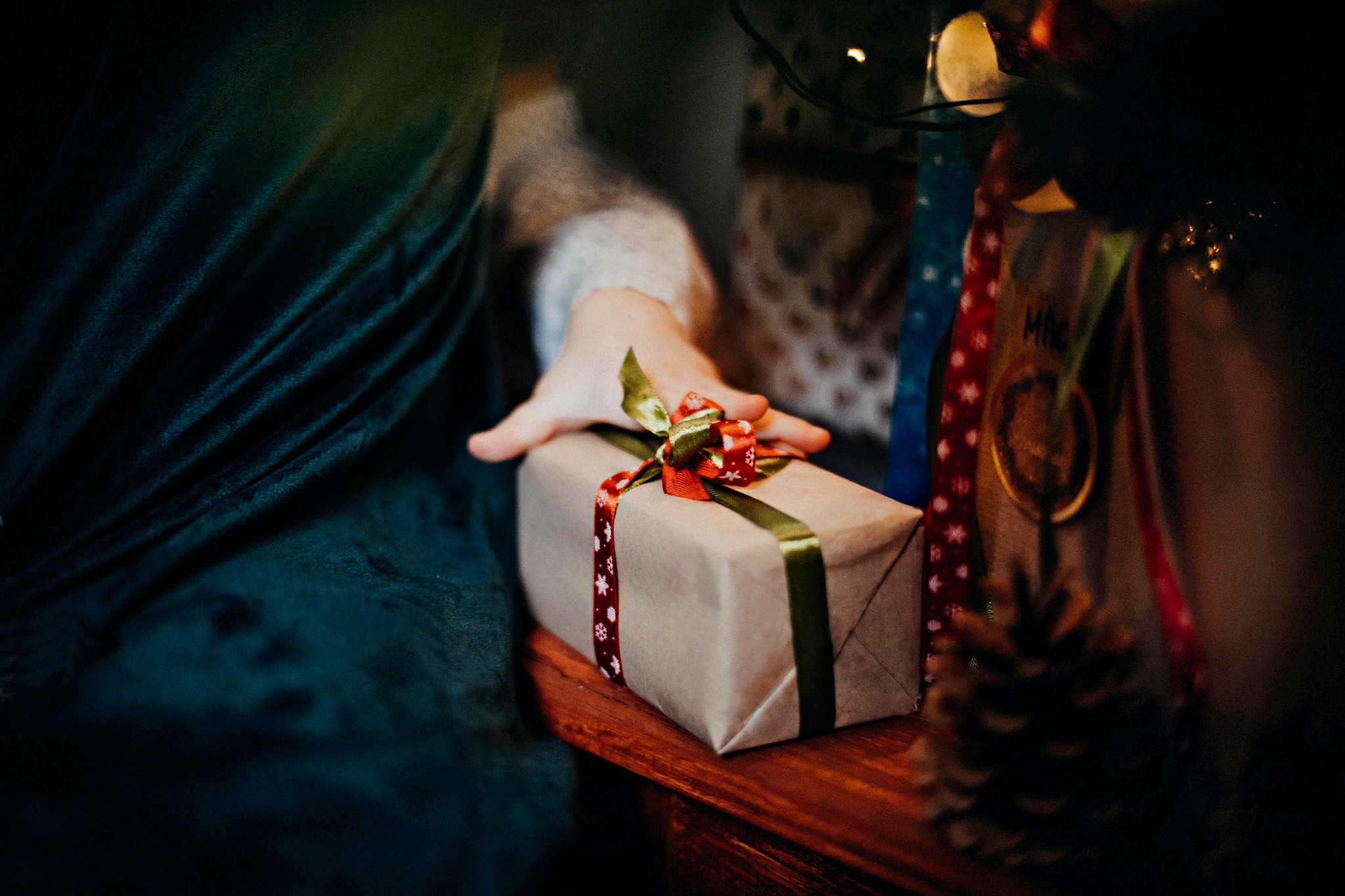 Dłoń dziecka sięga po prezent zapakowany w szary papier i przewiązany zieloną oraz czerwoną wstążką w śnieżynki, leżący na drewnianym stole, w tle fragment zielonego aksamitnego materiału...