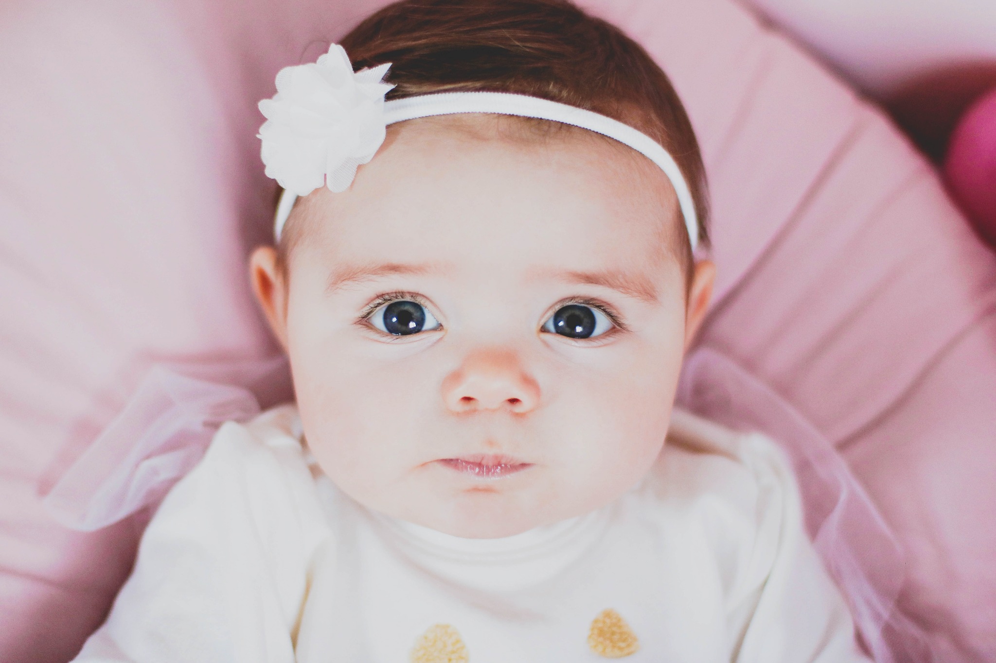 Portret niemowlęcia z ciemnymi oczami i białym kwiatem we włosach, ubrane w białą koszulkę z żółtymi aplikacjami na tle różowej tkaniny.