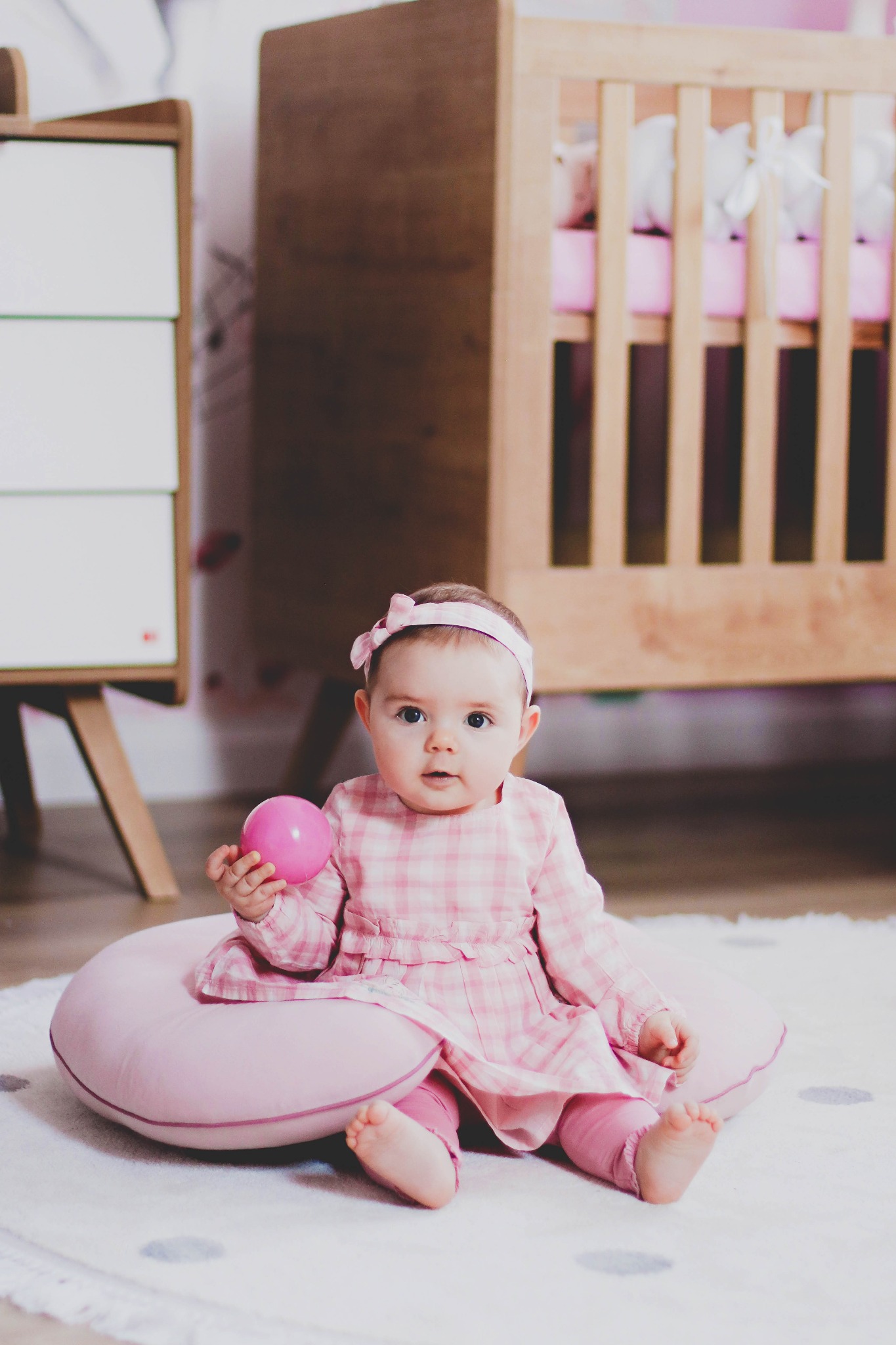 Urocza dziewczynka w różowej sukience w kratkę i opasce z kokardką siedzi na różowej poduszce, trzymając różową piłkę na tle drewnianego łóżeczka i białej komody.
