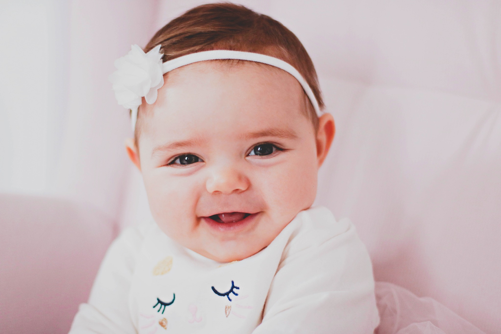 Uśmiechnięta niemowlęca dziewczynka z białym kwiatem na opasce, ubrana w białą bluzkę z haftowanymi oczami i złotymi elementami, na tle różowej poduszki.