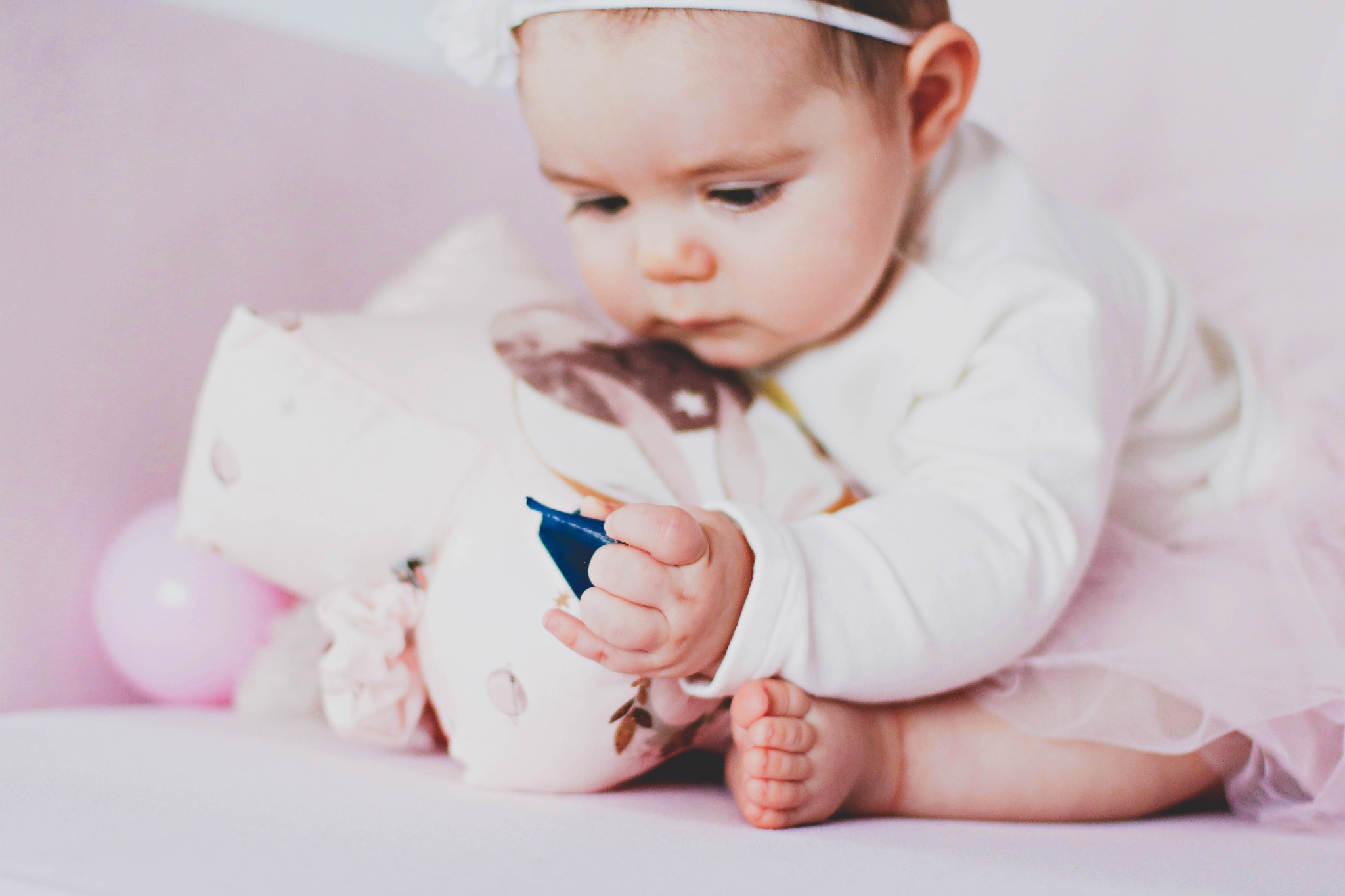 Niemowlę w tiulowej spódniczce i opasce z kokardką, siedzące na różowym tle, skupione na zabawie z pluszową zabawką i małym niebieskim przedmiotem w dłoni.