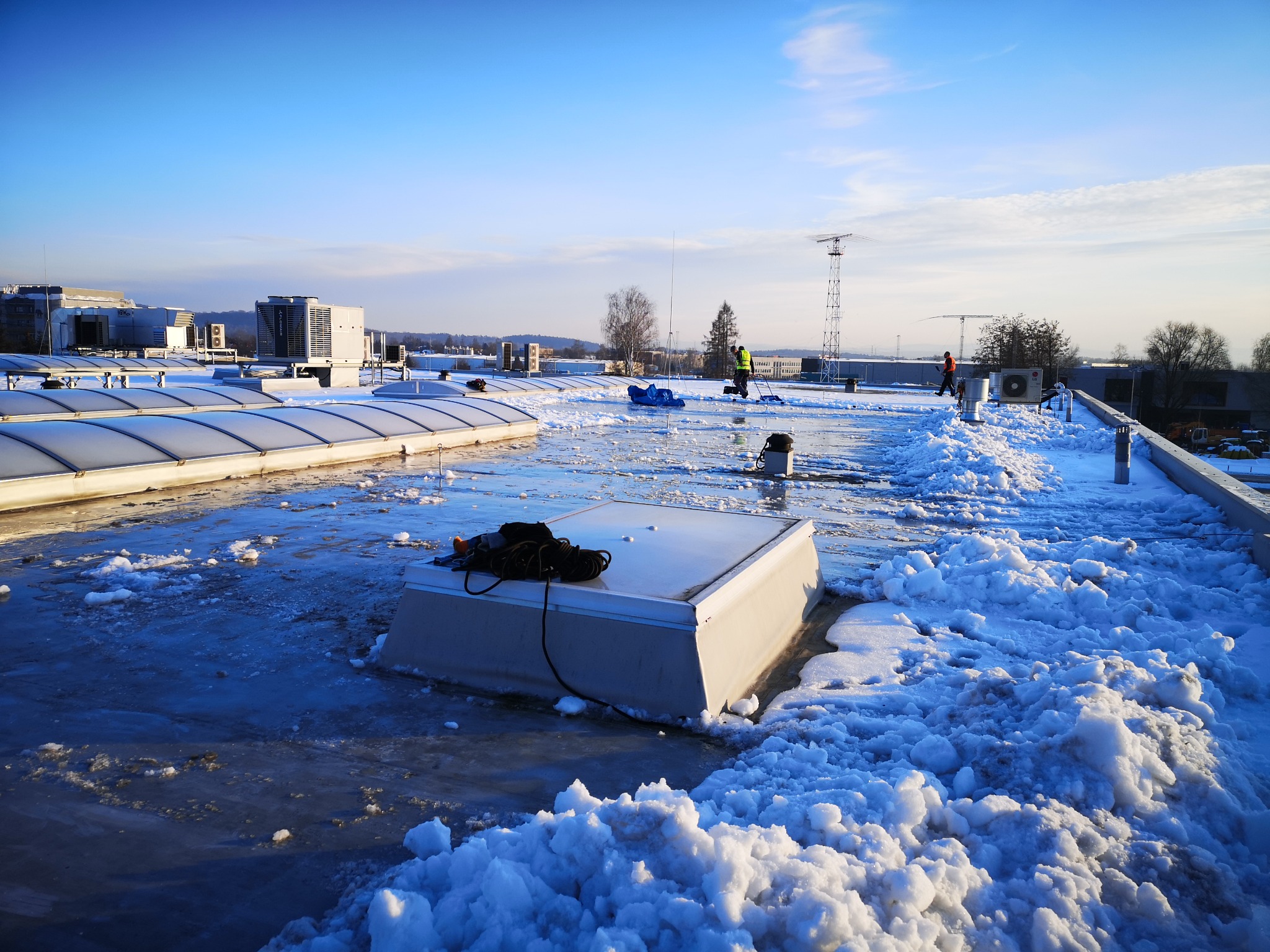 Pracownik w kamizelce odblaskowej odśnieża płaski dach budynku w Krakowie, na dachu leży śnieg i lód, widoczne świetliki dachowe i kominy wentylacyjne.