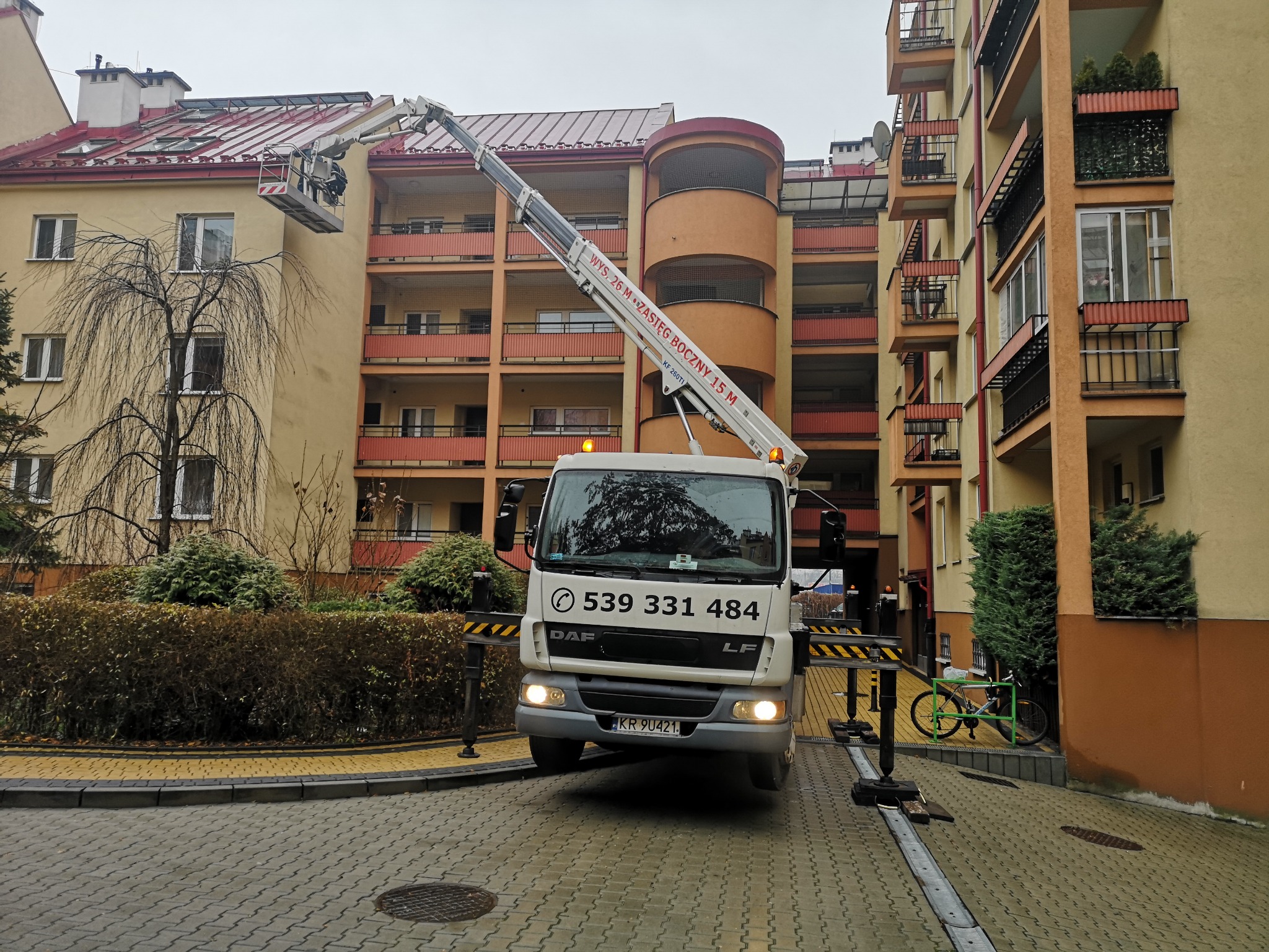 Podnośnik koszowy DAF na tle wielopiętrowego budynku, z wysięgnikiem sięgającym dachu, prawdopodobnie podczas prac konserwacyjnych lub naprawczych.