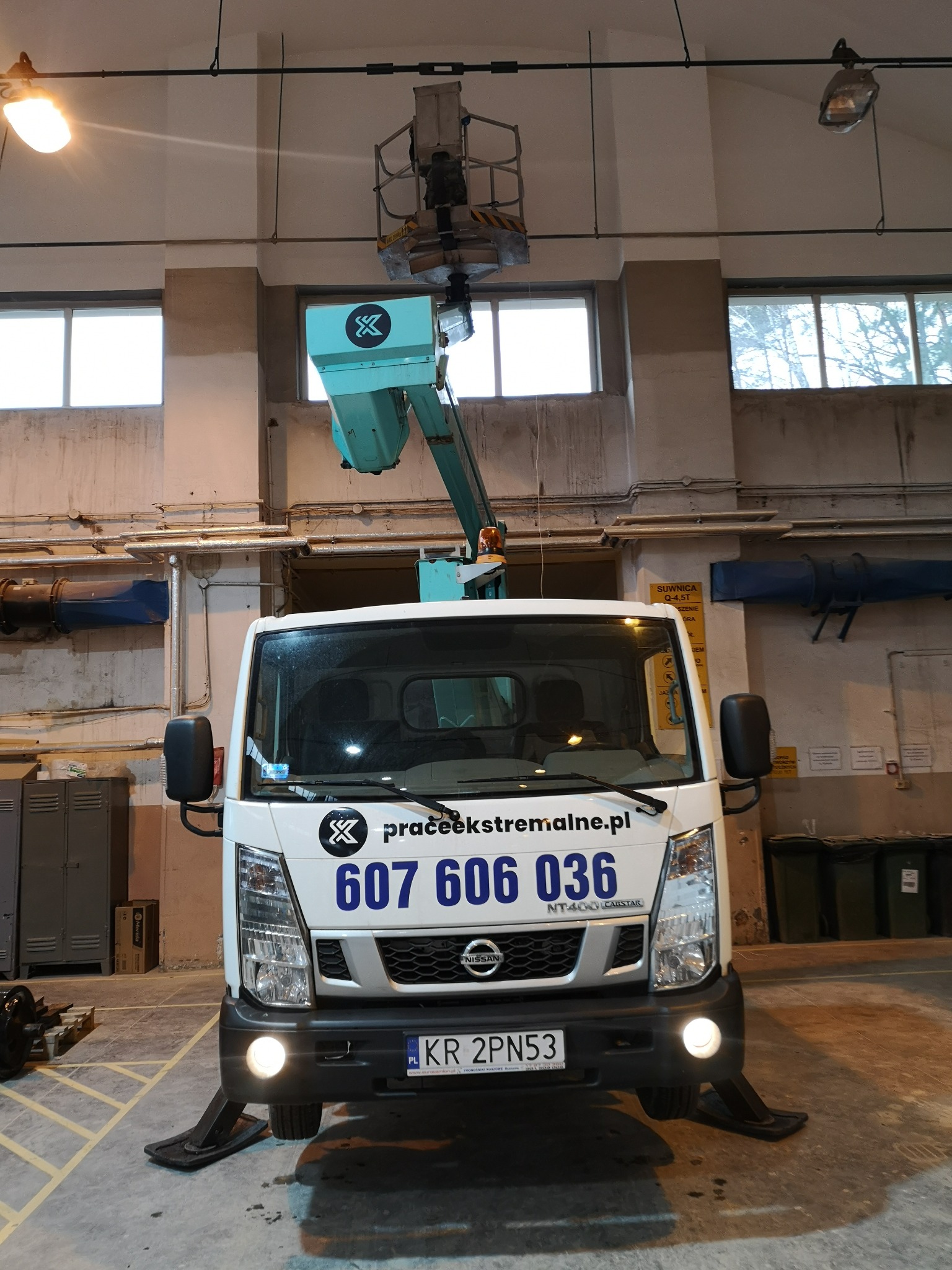 Podnośnik koszowy na białym samochodzie dostawczym z wysięgnikiem ustawiony w hali, widoczne podpory stabilizujące pojazd, logo firmy na maszynie i numer telefonu na kabinie.
