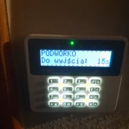 Panel sterowania alarmem Satel Perfecta z podświetlanymi przyciskami, wyświetlający komunikat 'PODWORKO Do wyjścia: 15s' i świecącą się zieloną diodą.