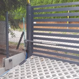 EZ INSTAL - Szara, pozioma brama z automatycznym napędem, widok na kostkę ażurową i ogrodzenie z siatki.