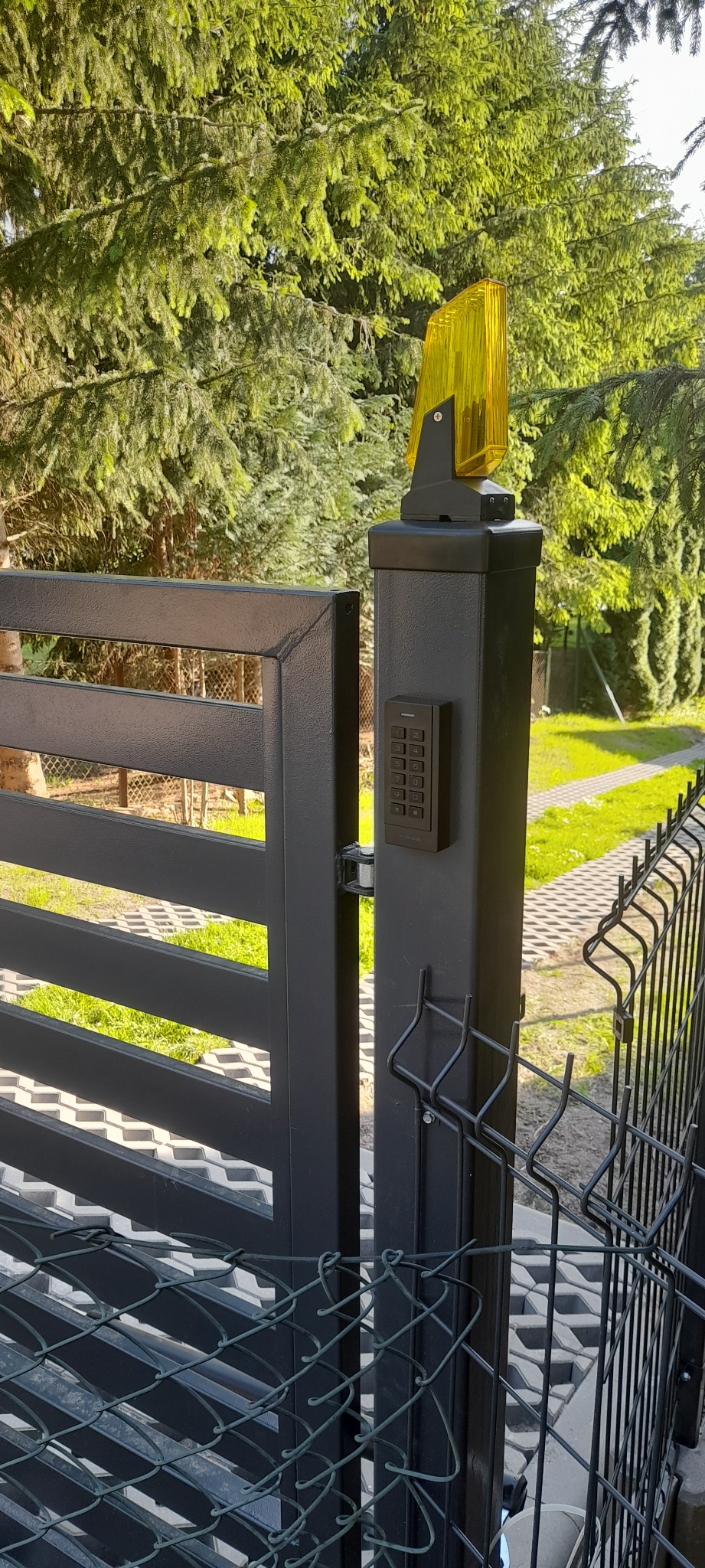 Słup bramy z domofonem cyfrowym, lampą ostrzegawczą i fragmentem ogrodzenia panelowego na tle zieleni.