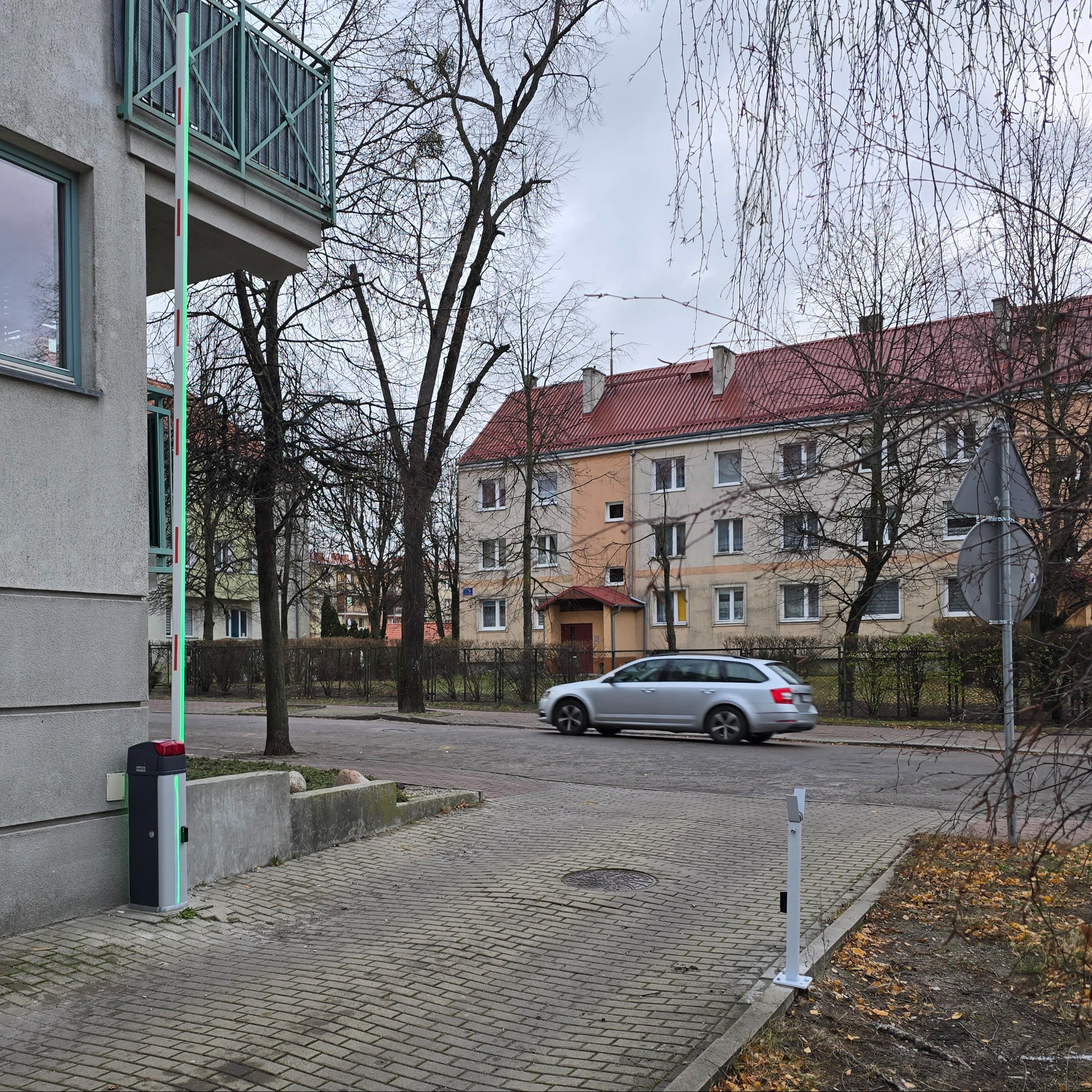 Szlaban automatyczny z podświetleniem LED przy wjeździe na osiedle, szary budynek w tle, przejeżdżający samochód.