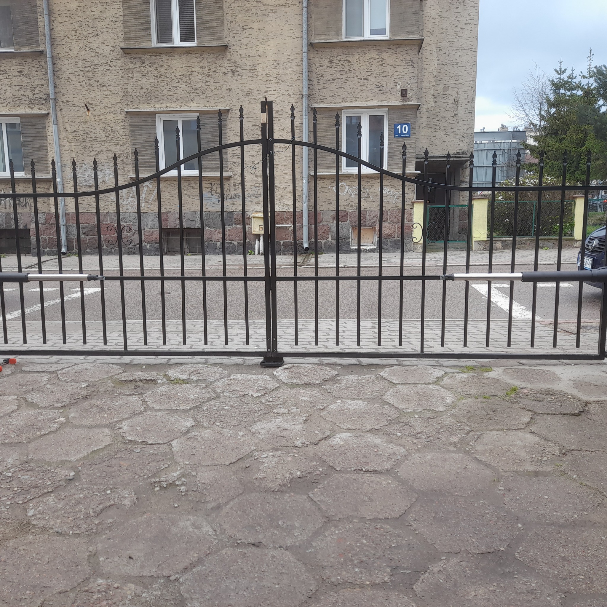 Czarna, metalowa brama wjazdowa z automatyką, widoczny siłownik, na tle budynku mieszkalnego z numerem 10.