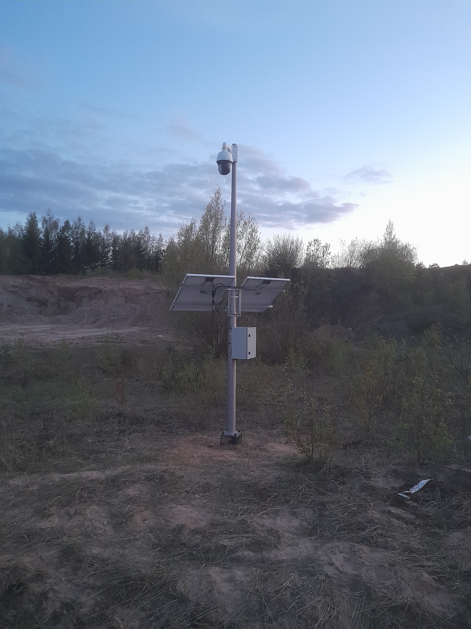 Kamera monitoringu na metalowym słupie z panelami słonecznymi, umieszczona na zewnątrz w terenie piaszczystym z niską roślinnością, pod wieczornym niebem.