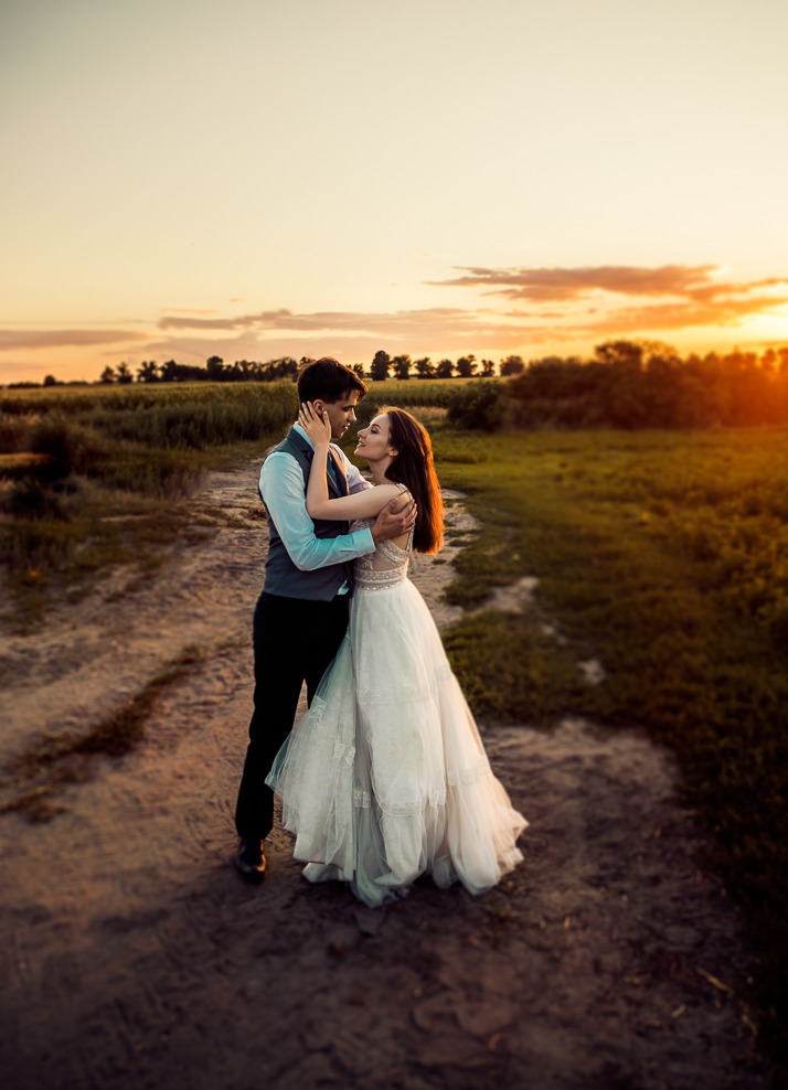 Para młoda w objęciach na polnej drodze o zachodzie słońca, panna młoda w koronkowej sukni, pan młody w kamizelce.