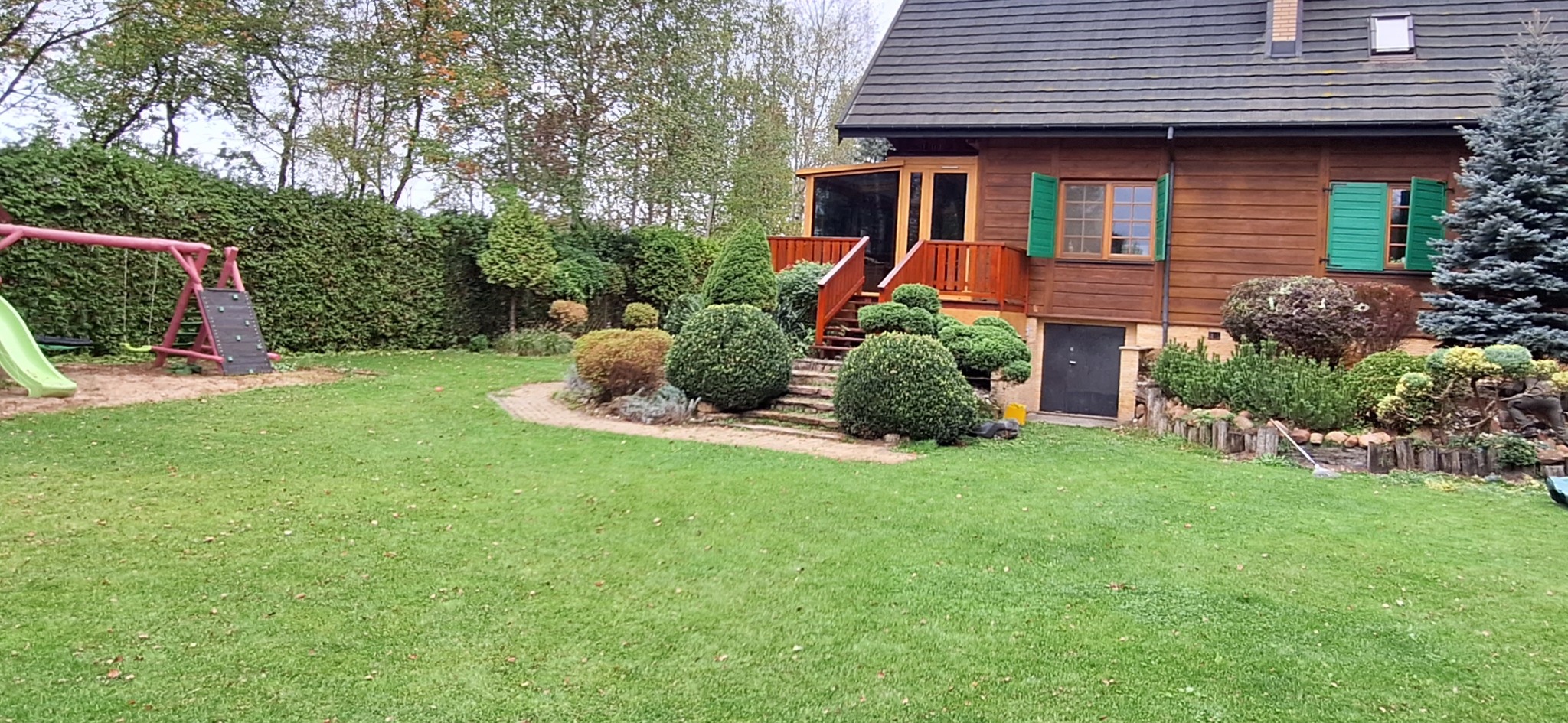 Utrzymany w dobrym stanie ogród z równo przystrzyżonym trawnikiem, ozdobnymi krzewami i drewnianym domem z zielonymi okiennicami w tle, widok z perspektywy trawnika.
