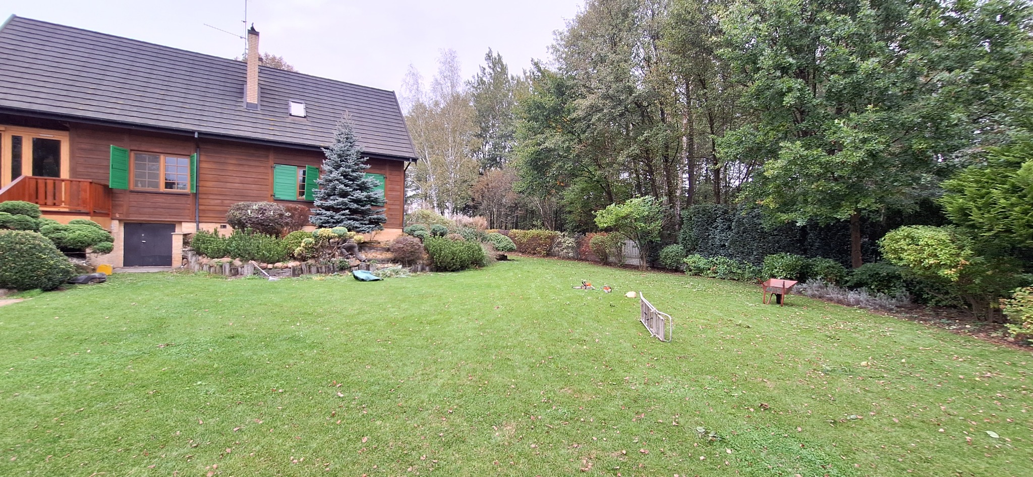 Rozległy trawnik przed drewnianym domem z zielonymi okiennicami, widoczne krzewy ozdobne, drzewa iglaste, drabina, taczka i dwie kosiarki spalinowe na trawie.