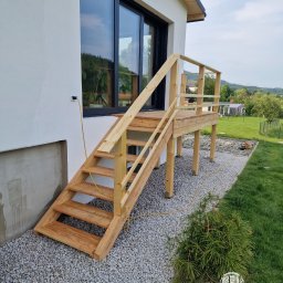 Paweł Śliwiński - Drewniane schody z balustradą prowadzące do drzwi balkonowych, z widocznym kablem zasilającym i elementami krajobrazu w tle. Jasne drewno, żwirowe podłoże.