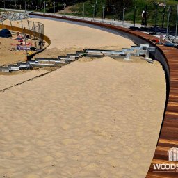 Paweł Śliwiński - Budowa drewnianego tarasu na plaży z metalowymi schodami i widocznym piaskiem. W tle osoby odpoczywające na plaży, całość ogrodzona siatką.