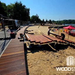 Paweł Śliwiński - Budowa drewnianego tarasu na plaży z widokiem na jezioro. W tle osoby odpoczywające na piasku, a w pierwszym planie pracownicy montujący konstrukcję.