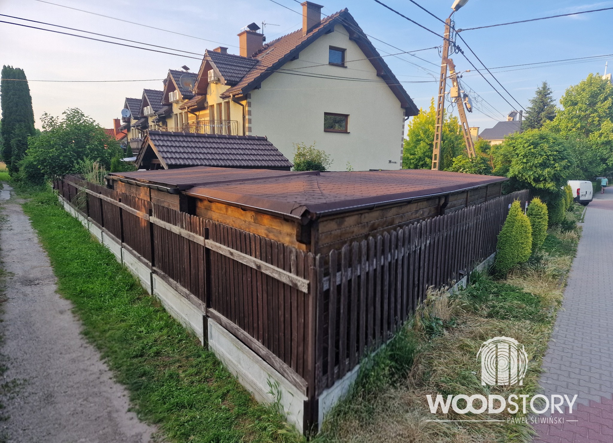 Drewniana konstrukcja z brązowym dachem, prawdopodobnie zadaszenie lub wiata, otoczona płotem z desek. W tle domy jednorodzinne i słup energetyczny.